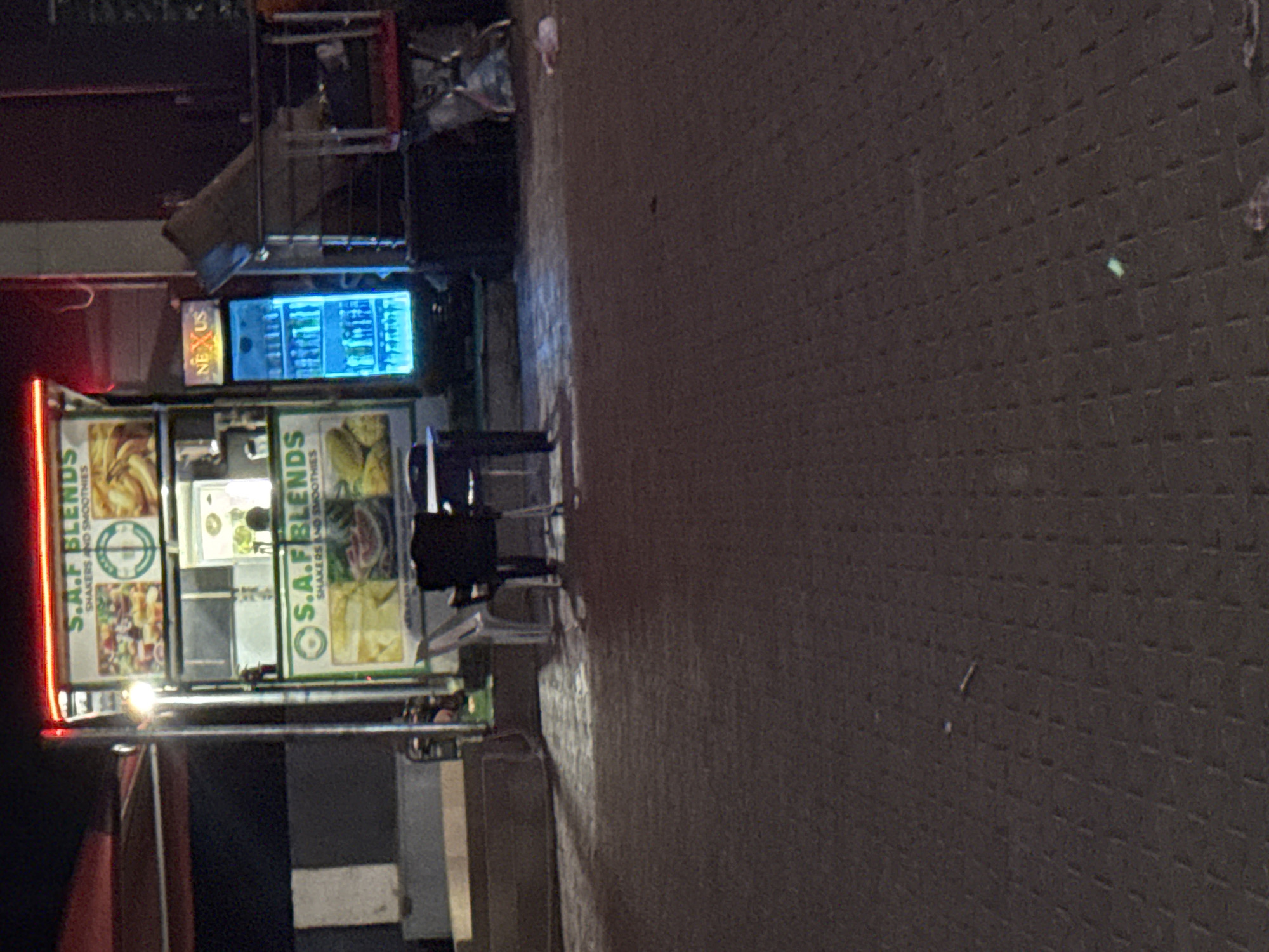 The image depicts a roadside smoothie stand in Kurunduma, Nigeria. The stand, named "S.A.F BLENDS", has an illuminated sign and displays images of its offerings. A brightly lit cooler with the name "NEXUS" on top is positioned nearby. Two people are seated at a small table in front of the stand. The stand is situated on a paved surface.