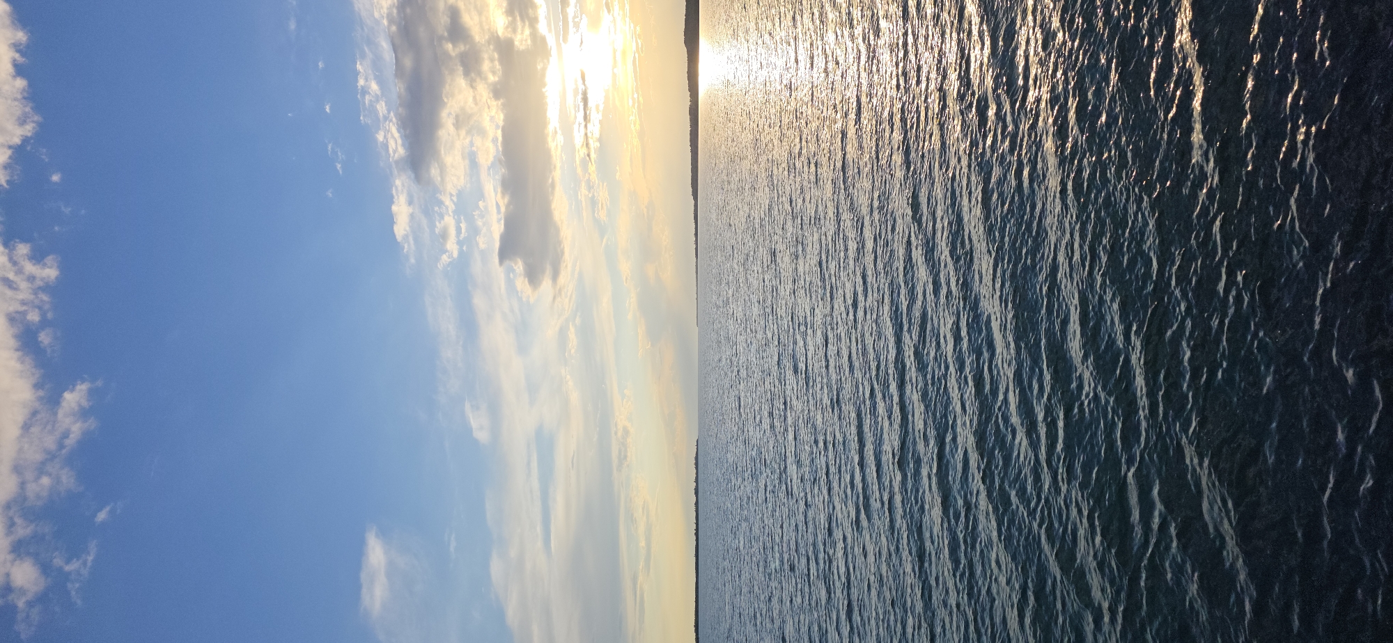 This is a vertical, eye-level shot of a calm body of water meeting the sky at sunset. The image is split diagonally, with the left side dominated by a bright blue sky filled with wispy white and grey clouds, some illuminated by the sun. The sun itself is a bright, hazy orb positioned just above the horizon on the right side of the frame, casting a warm, golden light that blends into the sky and reflects off the water.
The right side of the image is the water, likely a lake or sea, reflecting the sunset. The water's surface is rippled, creating a textured, shimmering effect as the sunlight catches the waves. The horizon line is clearly visible, separating the sky and water, with a faint, dark line of land visible in the distance. The overall mood is serene and peaceful, conveying the beauty of a natural landscape at dusk. The lighting suggests it is late afternoon or early evening. There is no visible text in the image.