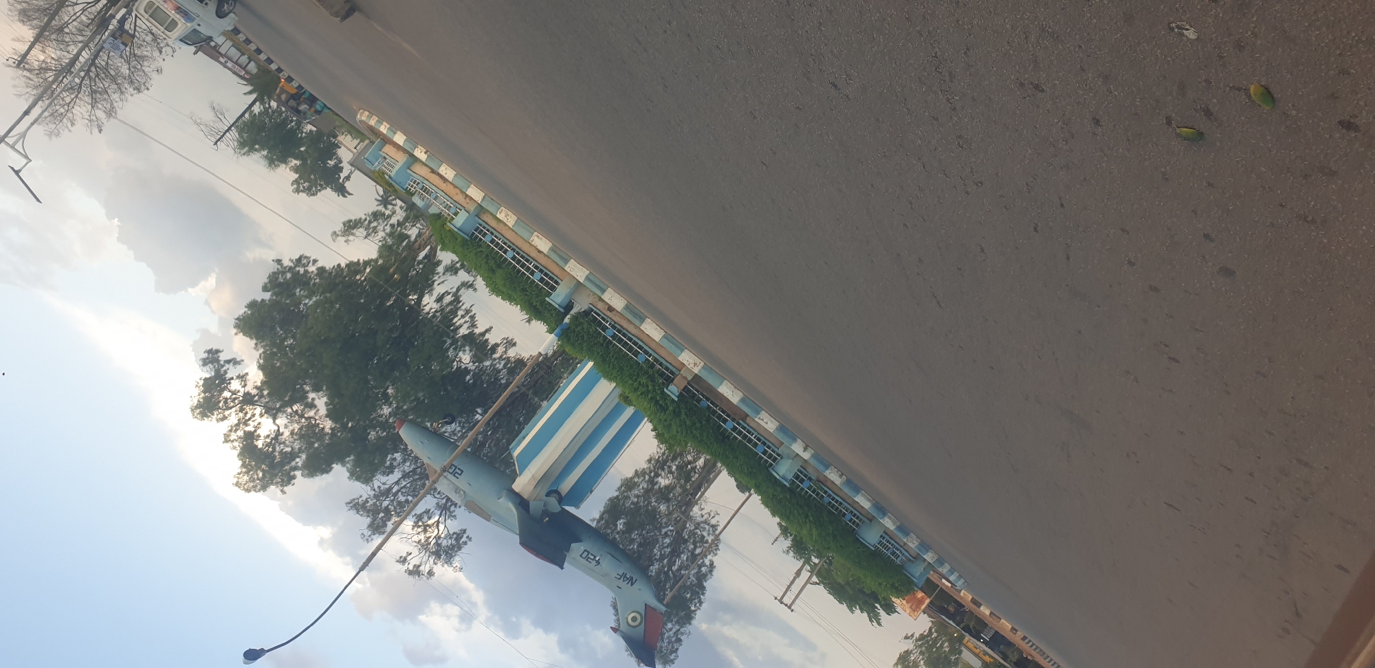 A light blue jet aircraft is displayed on a pedestal in the city of Barakin Lerawi, Nigeria. The aircraft is positioned at an upward angle, with its nose pointing towards the sky. It is mounted on a cylindrical pedestal that is painted with alternating blue and white stripes. The pedestal is surrounded by a low green hedge and a decorative fence with blue and white striped borders. The aircraft has markings on its tail, including a circular insignia with red, green, and white colors. The registration number "NAF 420" is visible on the fuselage. The scene is captured on a road, with asphalt visible in the foreground and extending into the background. Trees with dense green foliage are scattered throughout the scene, and utility poles with wires are visible in the distance. The sky is partly cloudy, with patches of sunlight breaking through. In the background, a white vehicle and some buildings are partially visible, suggesting an urban or semi-urban setting. The overall impression is that of a monument or display, possibly located near an airport or military installation.