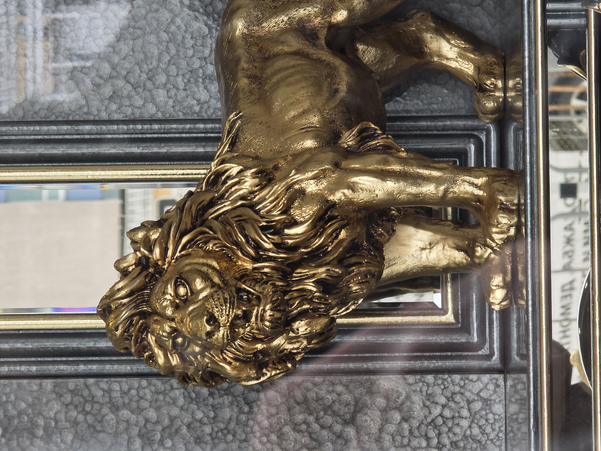 A golden lion sculpture is affixed to a grey textured surface with dark grey and gold trim. The sculpture is a three-dimensional depiction of a lion, with pronounced musculature and a detailed mane. Its head is visible in the lower left corner of the image, while its body extends upwards and to the right. The lion's paws appear to be gripping the surface to which it's attached. The background is a mix of textures, with reflective surfaces indicating the presence of glass or mirrors. The location is likely within a building or architectural structure in the city of Plovdiv, Bulgaria, given the decorative nature of the sculpture and its apparent integration into the building's design.