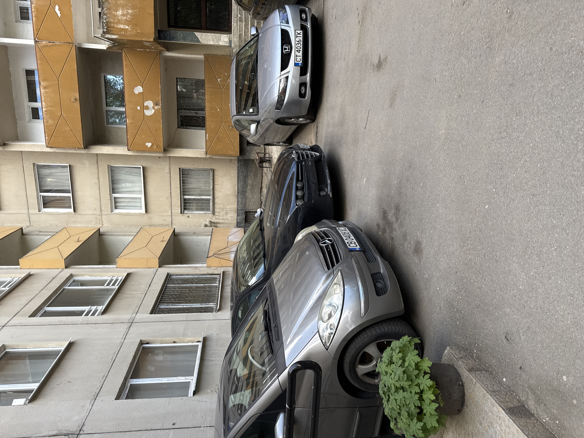 The image shows a street in Sofia, Bulgaria, with three parked cars in front of a building. The building is a multi-story residential building with a gray facade and yellow balconies. The cars are parked on a paved surface, and a potted plant is visible near the bottom right corner of the image. The car closest to the camera is gray, and its license plate reads "B 0409 BQ." Another car is dark colored, and the furthest car is silver, with a license plate reading "CT 4036 TK." The image is taken during the day, likely under natural light.