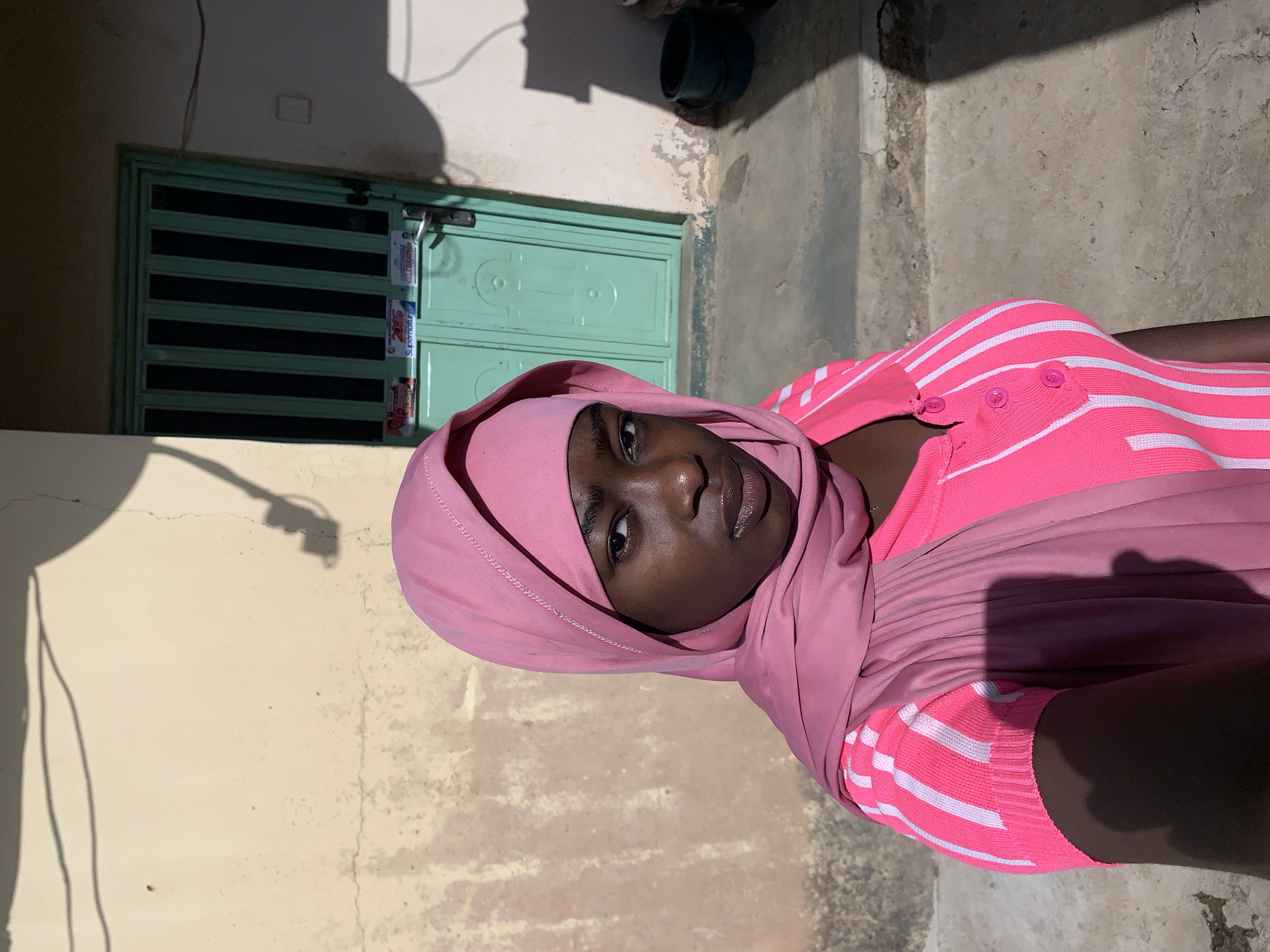 A woman wearing a pink headscarf and a pink and white striped top is centered in an outdoor shot. She is looking at the camera. Behind her, there is a light yellow wall on the left and a light green door with black bars and several stickers affixed to it. In the background, there is a concrete wall to the right and a dark object in the upper right corner. The scene is outdoors in what appears to be a courtyard. The location is Rafin Par, Nigeria.