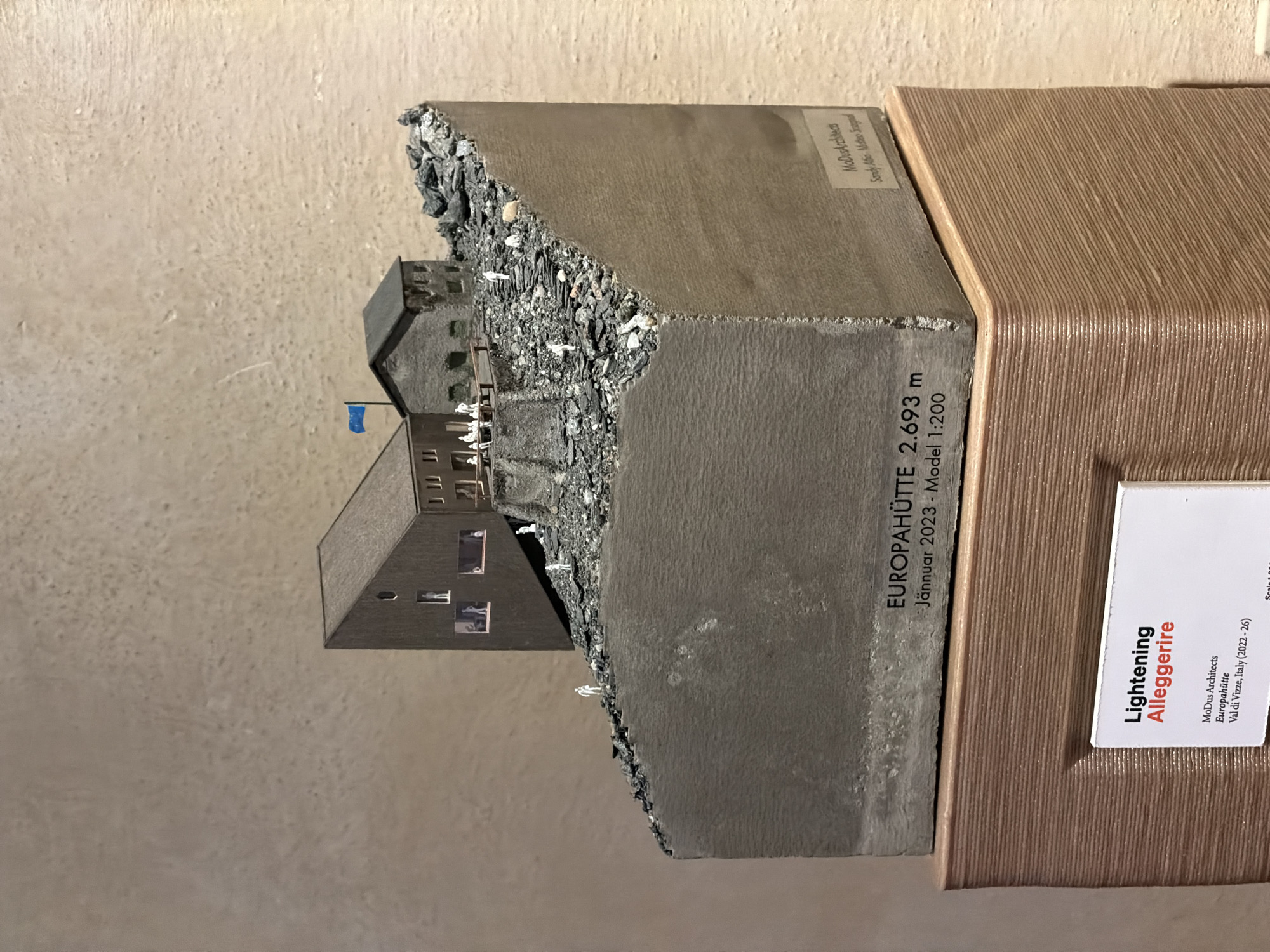 This image shows a model of the Europahütte, a mountain hut located at 2,693 meters above sea level. The model was created in January 2023 at a scale of 1:200. It sits on a display stand. A sign on the display stand indicates that the architects are MoDus Architects, and the project is called "Lightening Alleggerire." The hut is located in Val di Vizze, Italy and was built between 2022 and 2026.
