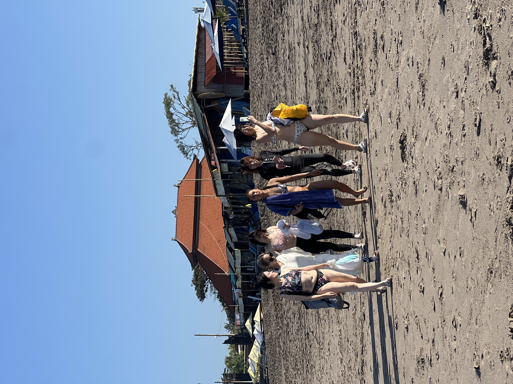 A group of six individuals are walking along a sandy beach in North Kuta, Indonesia. The individuals are all women and appear to be of East Asian descent. They are dressed in swimwear and some are carrying bags and towels. The beach is lined with umbrellas and lounge chairs, and there are buildings in the background, suggesting a resort or tourist area. The sky is clear and blue, and the sun is shining, indicating it is daytime. The sand is dark and wet in some areas, with footprints visible. The overall scene is one of leisure and recreation, with people enjoying a day at the beach.