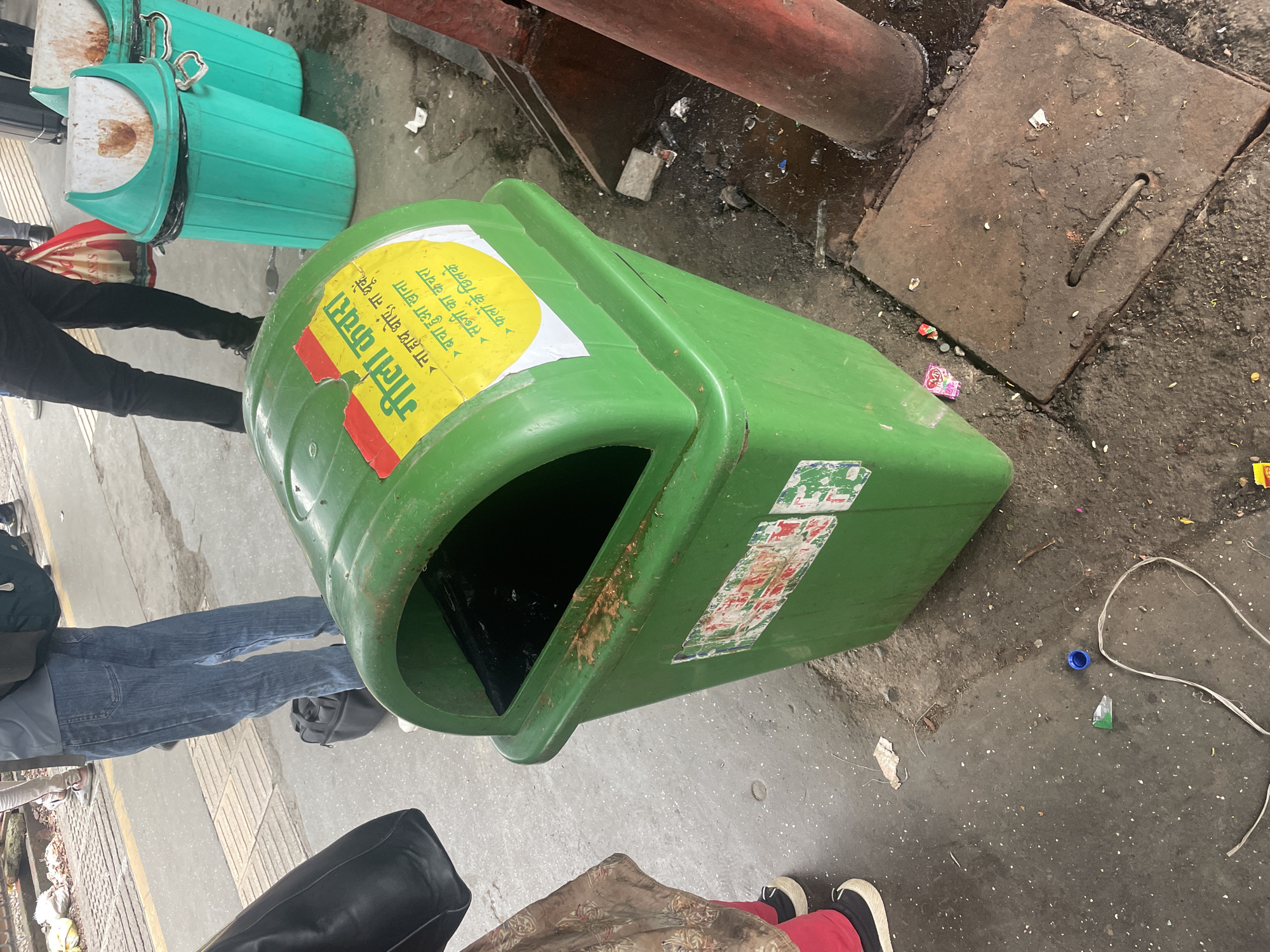 The image depicts a public space, likely a train platform in Mumbai, India, with a green waste bin prominently featured in the foreground. The bin has a yellow sticker with Hindi text and bullet points, indicating instructions for wet waste. The bin is filled with dark material, and there are various other bins, some green and some turquoise, visible behind it. The ground is concrete and appears wet, with scattered debris and a metal grate in the lower right quadrant. A portion of a person's legs, wearing blue jeans and dark shoes, is visible on the left, and another person's legs wearing black pants and dark shoes are in the background. A black bag is also visible on the ground near the person in jeans. The overall scene suggests a busy public area with a focus on waste management.