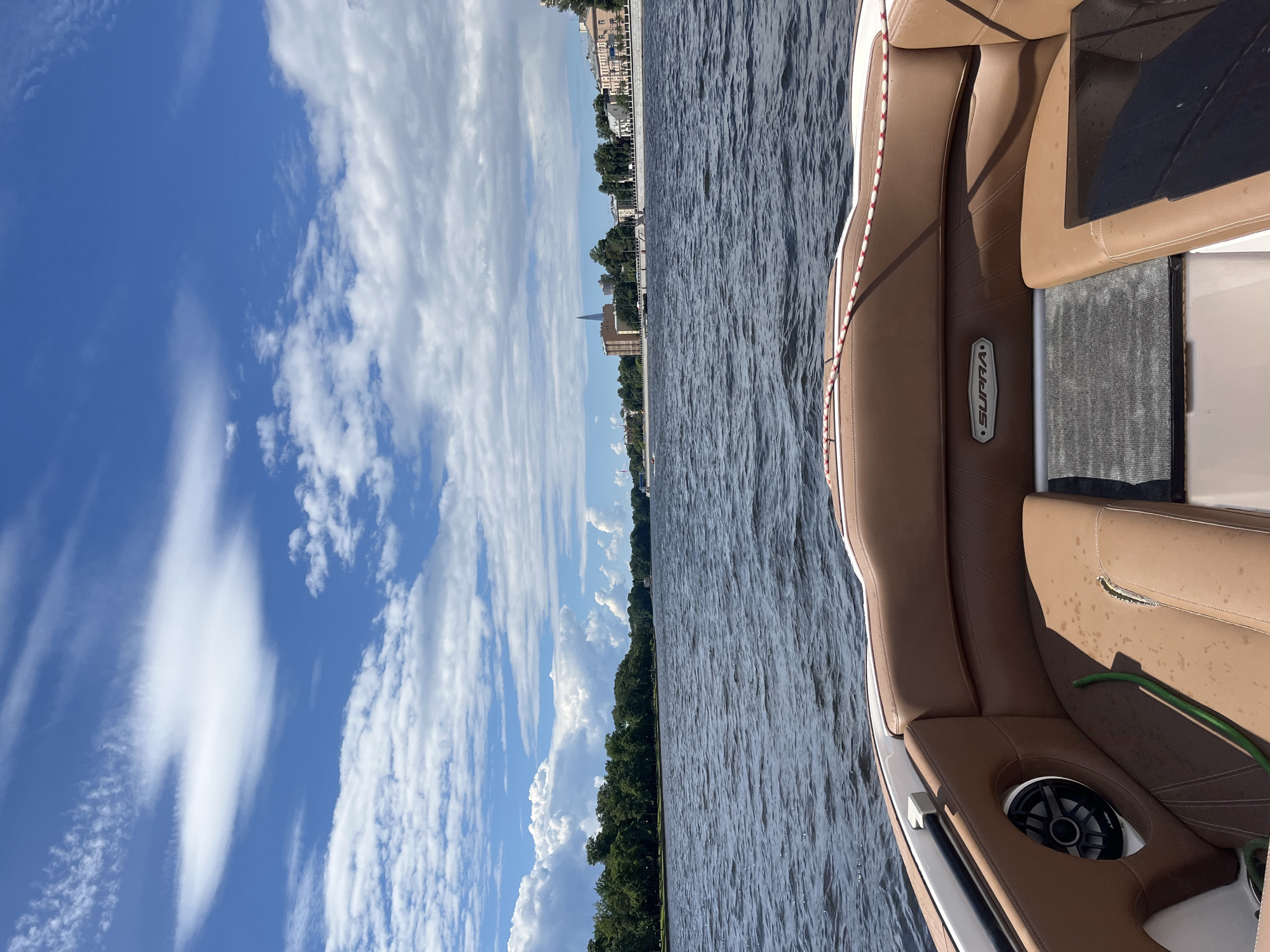 The image shows a boat on the water, with a view of the Saint Petersburg skyline in the distance. The boat has brown leather seats and a speaker visible. The water is calm with small waves. The sky is blue with white clouds. The skyline features buildings and trees along the waterfront. There is text on the boat that reads "SUPRA". The scene appears to be during the daytime, with clear weather. The overall mood is peaceful and scenic.