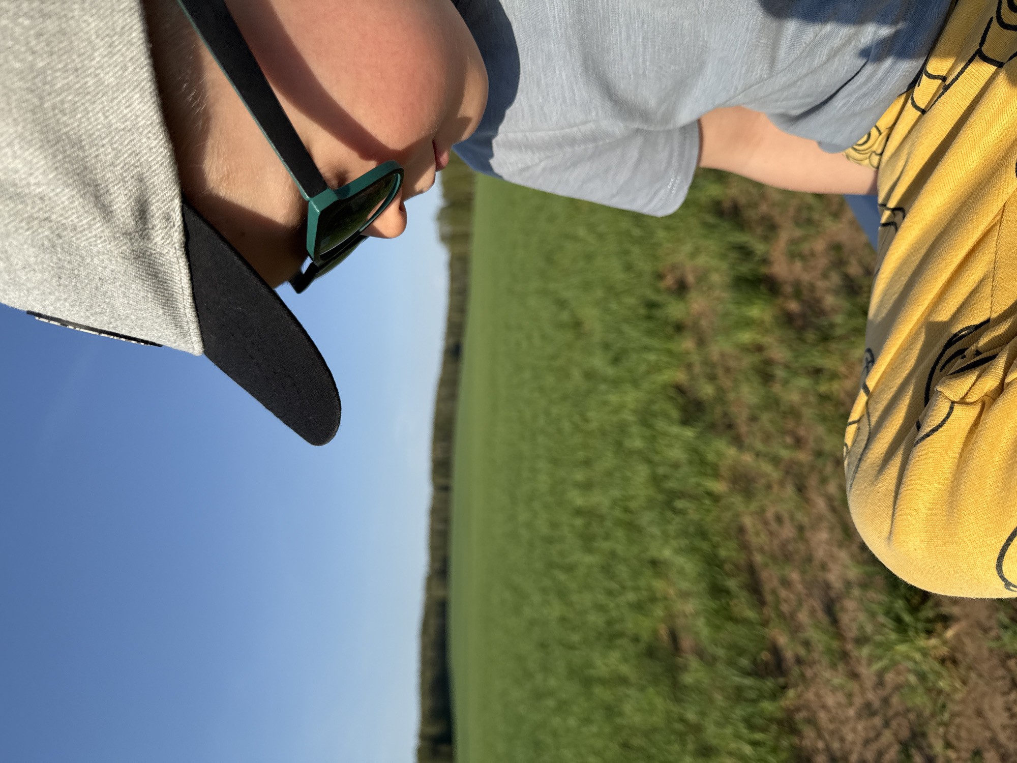 A child wearing a gray baseball cap and green-framed sunglasses is positioned on the left side of the image. The child has a light blue shirt and is wearing yellow pants. The background is a rural scene with a green field and a clear blue sky, suggesting a daytime outdoor setting, in the city of Шумиха, Russia.