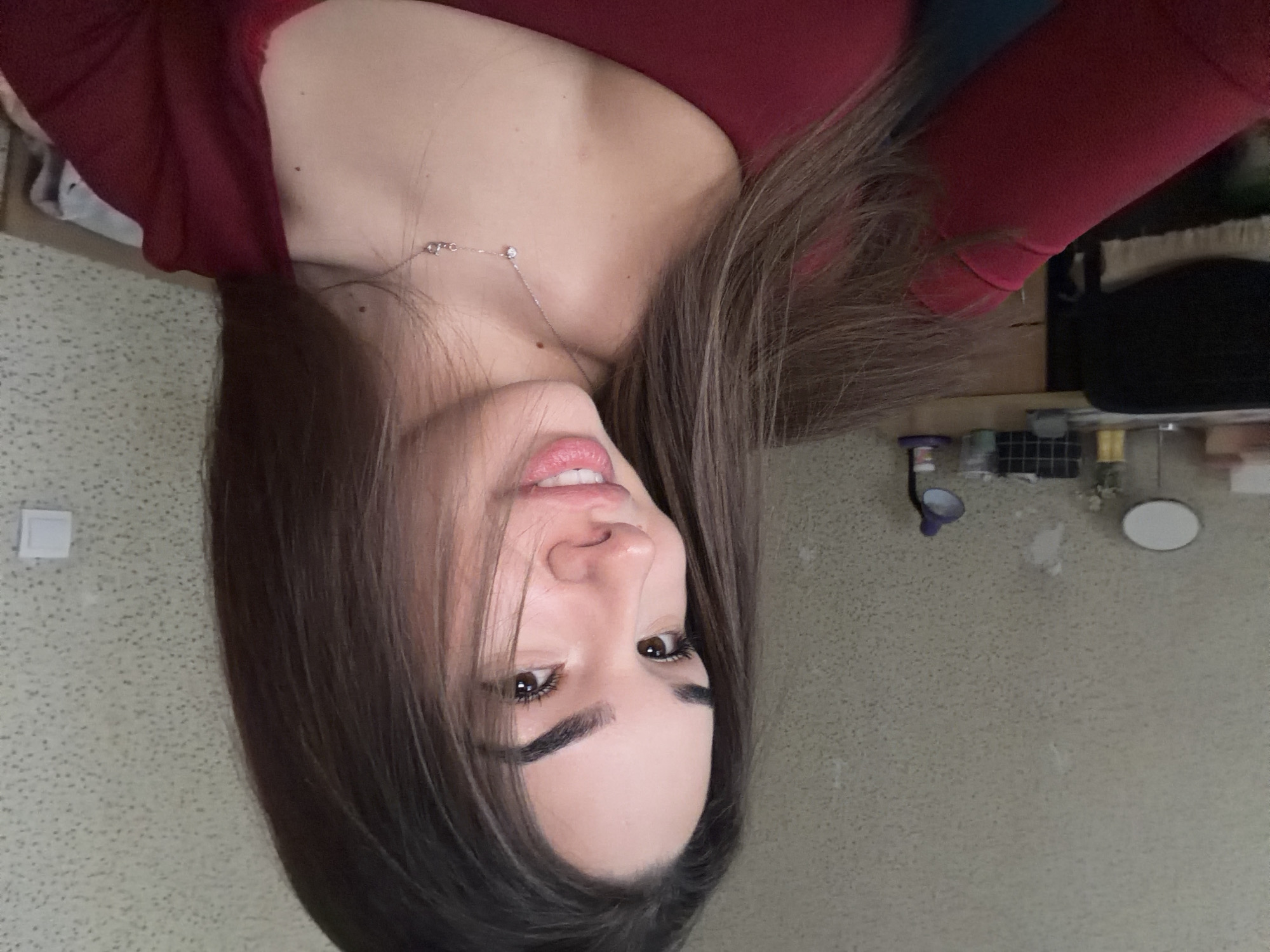 A young adult female is centered in a medium close-up, posing indoors in what is identified as Sofia, Bulgaria. She has long, dark brown hair, parted to the right, which partially obscures the left side of her face. Her complexion is light, and she has dark brown eyes, arched eyebrows, and light pink lips formed into a subtle smile, revealing white teeth. She is wearing a solid red, long-sleeved top with a wide, rounded neckline. Around her neck is a delicate silver-colored chain necklace with two small, clear adornments, one appearing heart-shaped and the other a small bar with an inscription.
The background consists of a textured, light beige wallpaper with a fine, speckled pattern. To the left, a dark office chair is partially visible, along with a light-colored wooden desk. On the desk, from left to right, are a white oval-shaped mirror on a stand, several small unidentified containers, a small vase with faint floral elements, a grid-patterned organizer containing various items, and a purple-hooded desk lamp with a black stand. To the right, on the wall, a white square electrical outlet is mounted. The lighting is even, suggesting an indoor ambient light source.