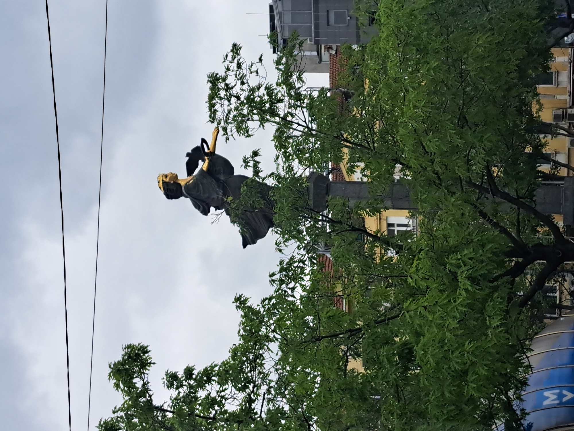 The image captures a statue atop a column in Sofia, Bulgaria, framed by a tree with green foliage. The sky is overcast. Power lines are visible in the upper left corner of the image. Behind the tree, a building with a yellow facade and white windows can be seen. Another building with a gray exterior is also visible in the background. The base of a blue object with a white Σ symbol can be seen in the bottom right corner. A bird sits atop the statue's head.