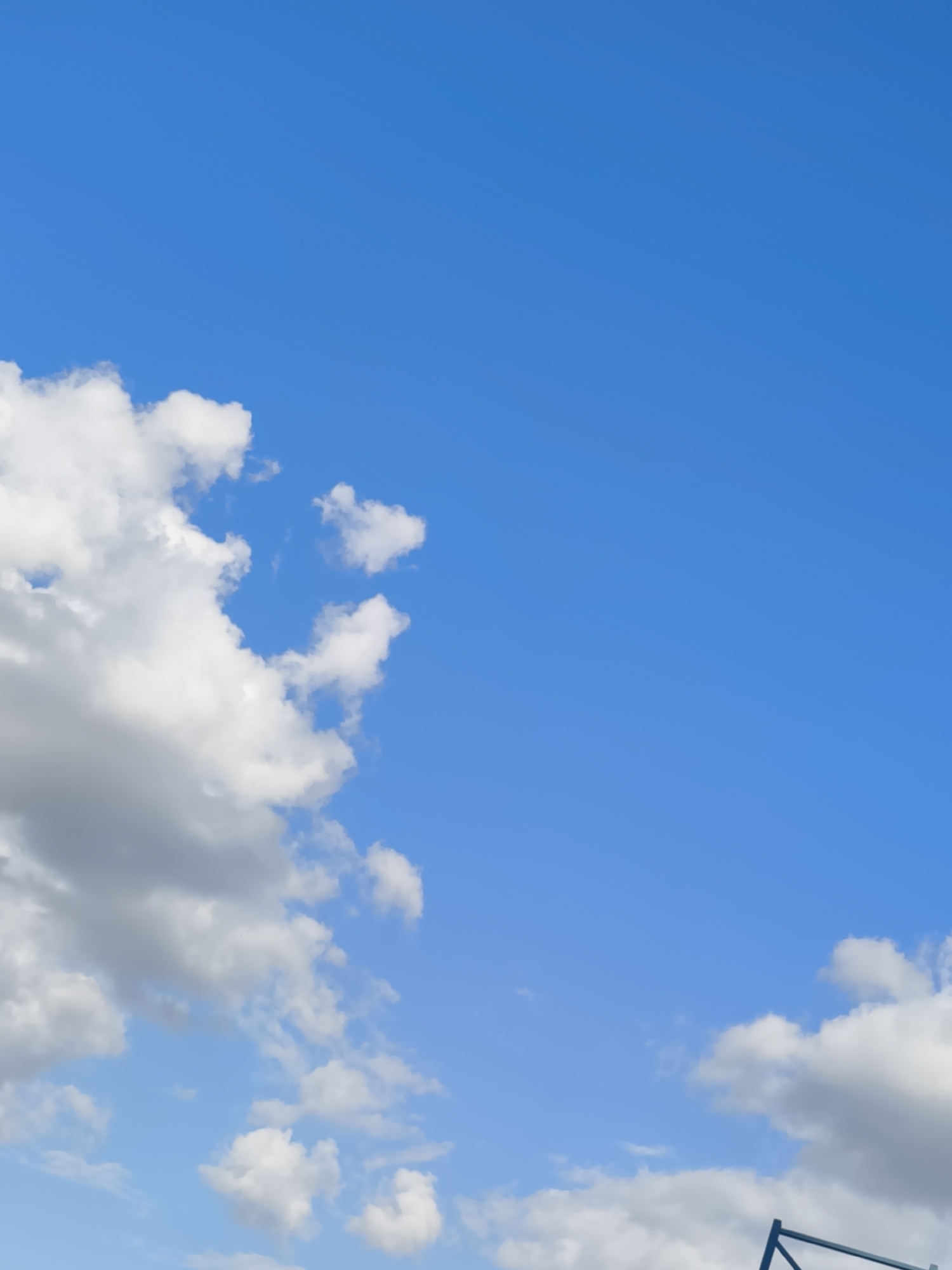 The image depicts a clear blue sky with scattered white and gray cumulus clouds. The lower portion of the image shows a dark blue metal structure, possibly part of a construction or an outdoor fixture. The scene appears to be an outdoor environment, likely a park or an open area within Chișinău, Moldova, given the information. No people are visible in the frame.