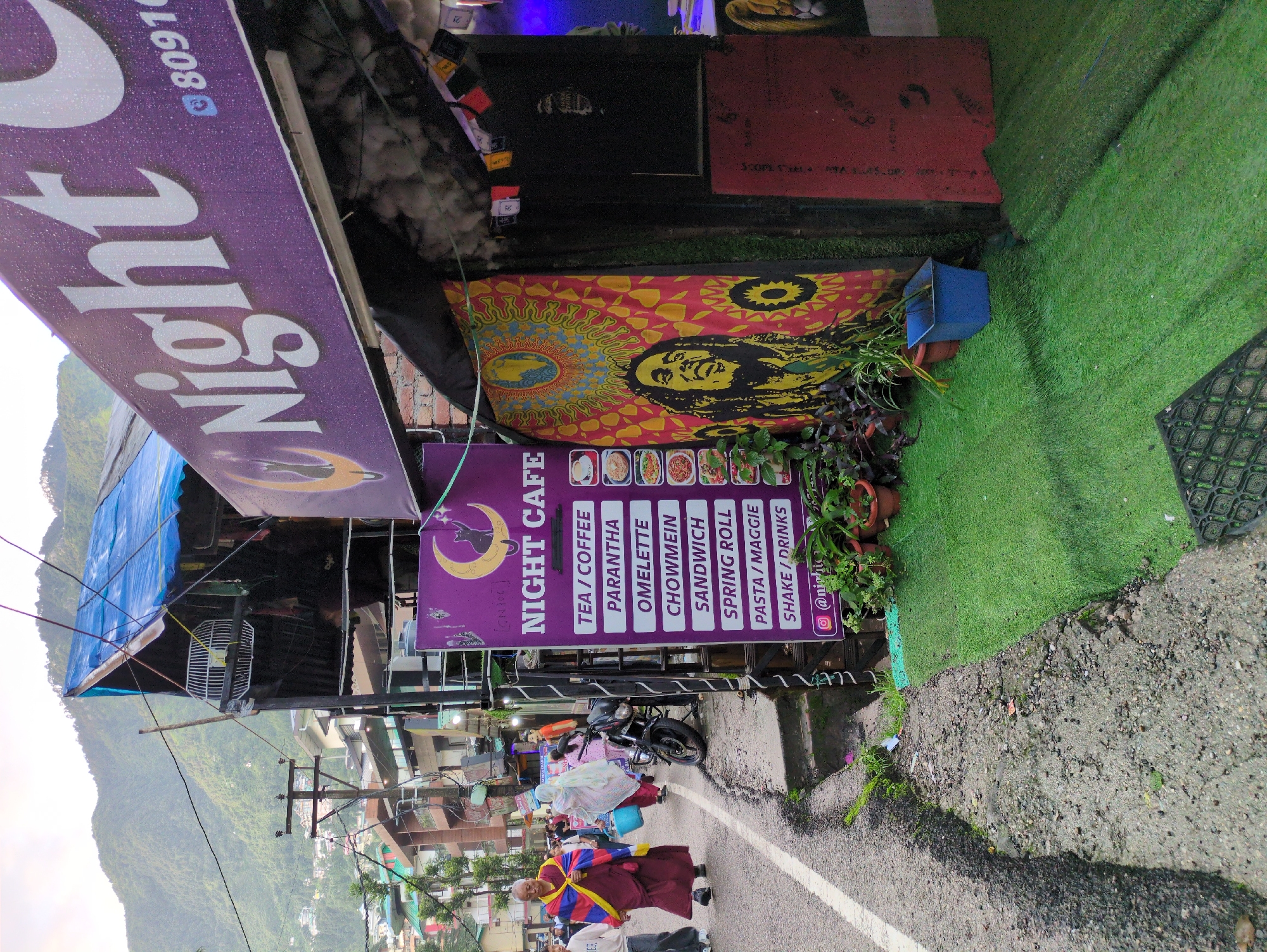 The image depicts a street scene in Bhagsu, India, featuring a cafe called "Night Cafe" with a prominent purple sign. The cafe appears to be a small establishment, possibly a street-side stall or a shop front. On the left side of the frame, a person dressed in traditional Tibetan attire, a maroon robe with blue and yellow stripes, is walking on the street. Other pedestrians are visible in the background. A motorcycle is parked near the cafe entrance.
The cafe's facade is decorated with a vibrant tapestry featuring a portrait of Bob Marley. A menu board is displayed vertically, listing items like "Tea/Coffee," "Parantha," "Omelette," "Chowmein," "Sandwich," "Spring Roll," "Pasta/Maggie," and "Shake/Drinks." The area in front of the cafe is covered with artificial green turf, with several potted plants adding to the decor. Small prayer flags are hung above the cafe entrance. The background shows a street lined with buildings and a backdrop of lush green hills, characteristic of the Bhagsu area. Power lines and a utility pole are visible, indicating the urban environment. The overall impression is a lively, casual atmosphere, typical of a tourist or local hangout spot in India.