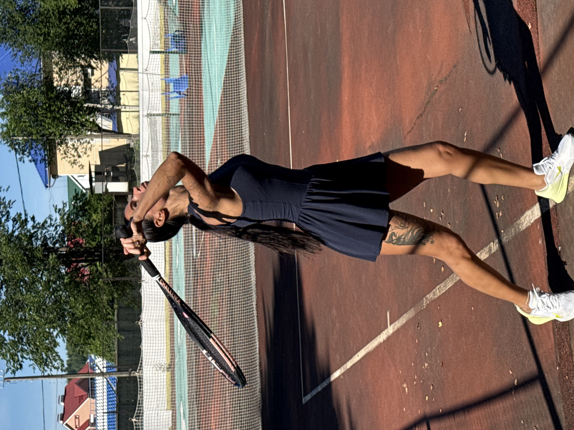 A woman is playing tennis on an outdoor court. She is wearing a dark blue tennis dress and white athletic shoes with neon yellow accents. She has long dark hair and a tattoo on her left thigh. She is holding a tennis racket above her head, in the process of serving. The court surface is red, with white lines marking the boundaries. In the background, there is a tennis net, trees, and buildings with red and blue roofs. The sky is clear and blue. The image was taken in Chaikovsky, Russia.