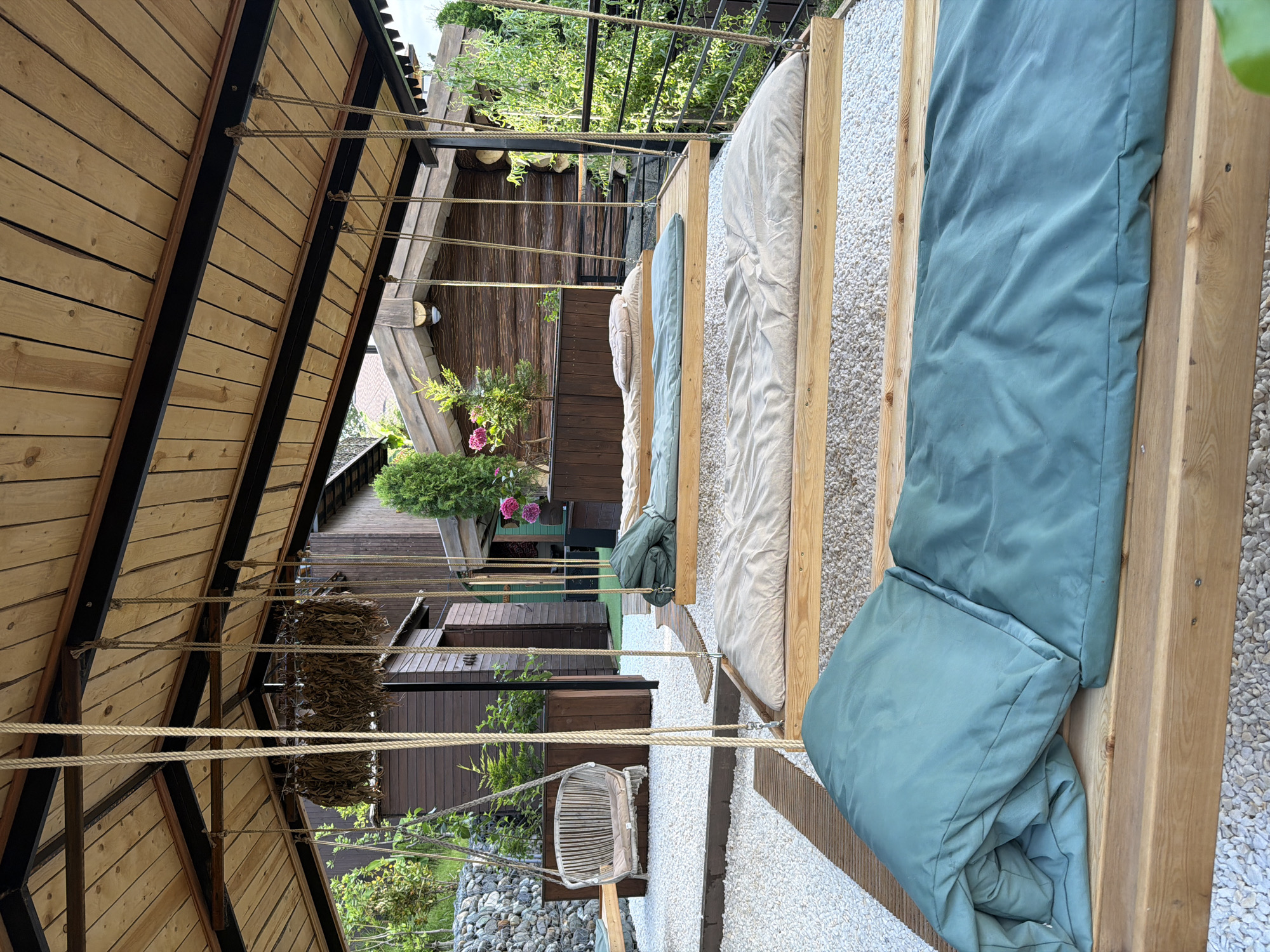 A covered outdoor area features several wooden swings with mattresses and pillows, set on a gravel ground. The swings are arranged in a row and are suspended by ropes from a wooden ceiling. In the background, there are wooden structures resembling small cabins and lush green trees. A stone wall and a wicker chair are also visible. The overall scene suggests a serene and natural outdoor space.