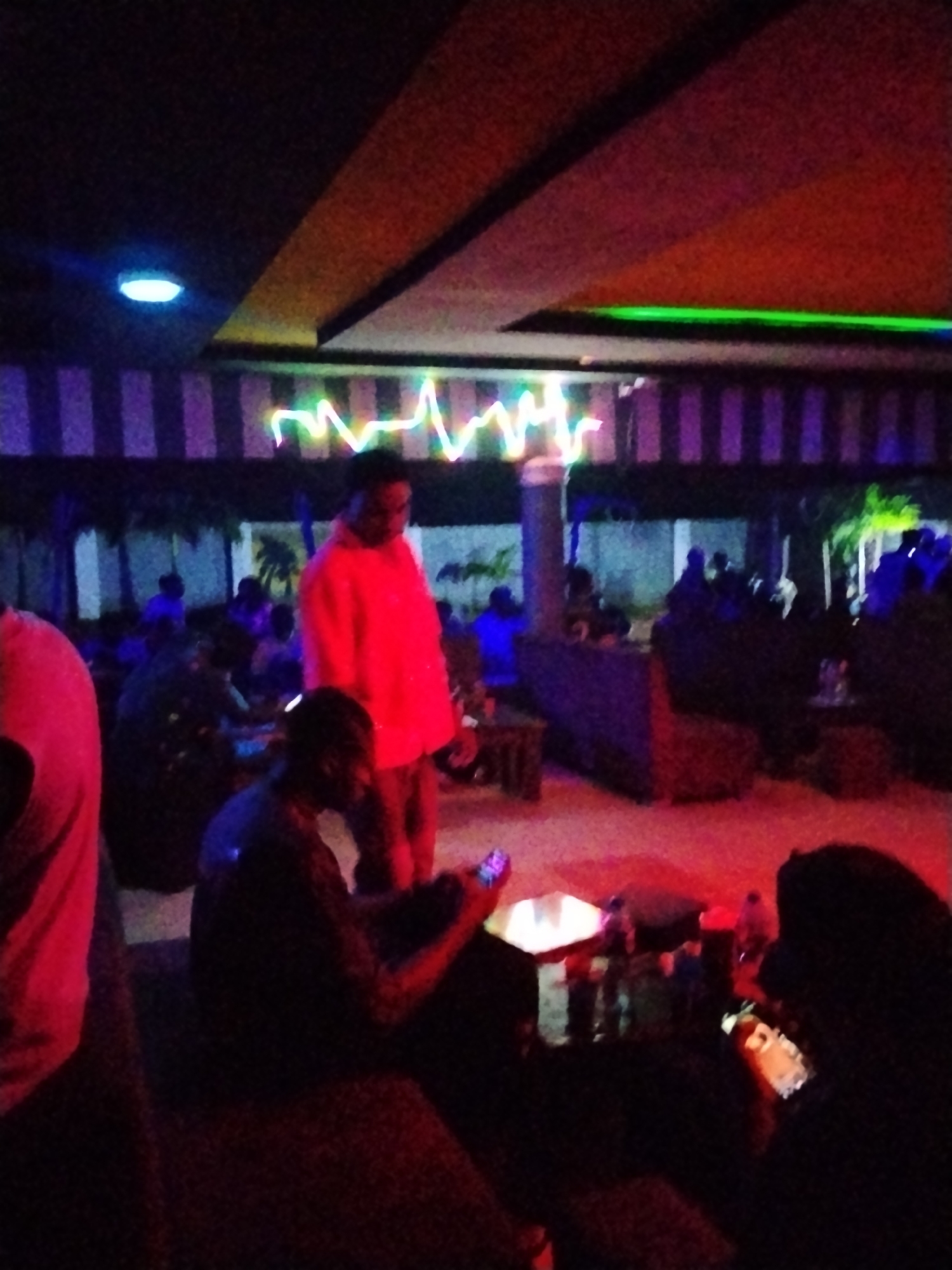 The image depicts an indoor gathering, possibly a club or lounge, illuminated by neon lights and ambient lighting in the city of Durimi, Nigeria. A person in a bright red jacket stands prominently in the foreground, facing away from the camera and towards a group of seated individuals. Several other figures are visible in the background, engaged in various activities within the dimly lit space. The environment features decorative striped walls and a textured ceiling with embedded lights. A table with items on it is discernible in the middle ground, suggesting a social or celebratory occasion. The overall atmosphere appears lively and social, characteristic of a night out in Durimi.