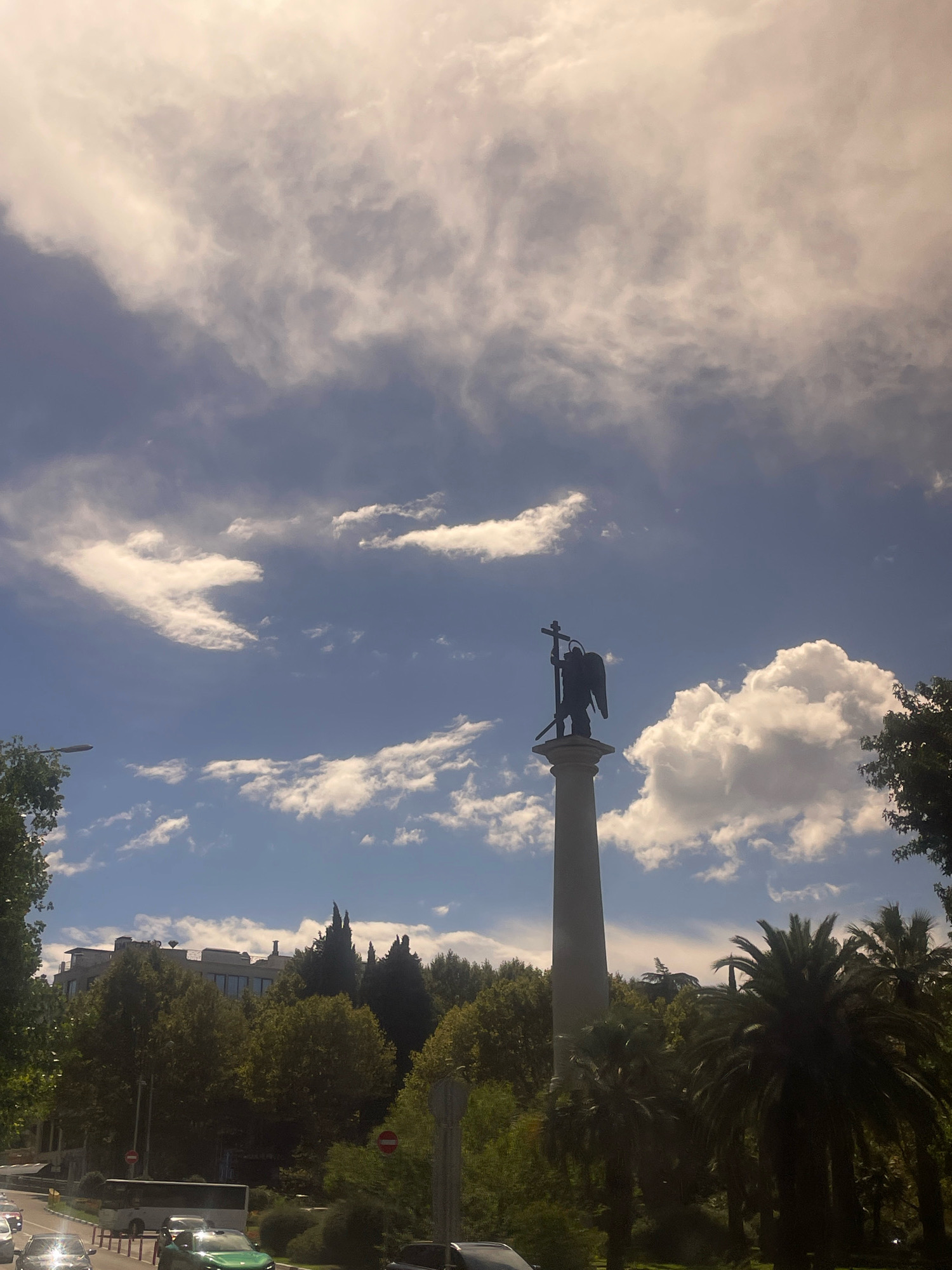 The image shows an angel statue on top of a column, silhouetted against a bright, cloudy sky. The statue holds a cross in its raised hand. In the foreground, there are trees, including palm trees, and a street with cars and a bus. Buildings are visible in the background, partially obscured by trees. The overall scene suggests an outdoor urban environment. The image is taken from a slightly low angle, looking up at the statue and the sky. The lighting suggests it is daytime, possibly late afternoon, with the sun filtering through the clouds.
The statue is located in Sochi, Russia. Specifically, it is the monument to the Archangel Michael, located at the beginning of Kurortny Prospekt. The statue is a symbol of the city and is a popular landmark.