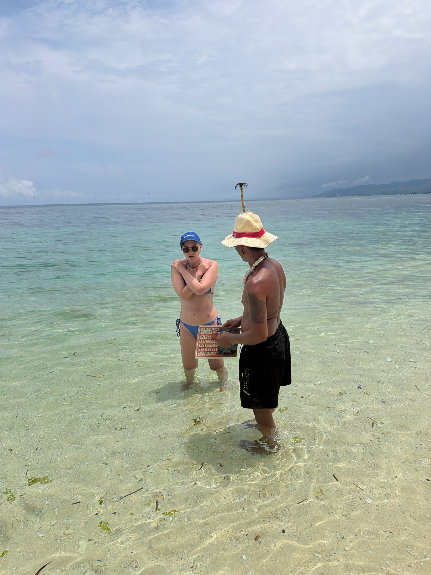 In the city of Malay, Philippines, a woman wearing a blue bikini and a blue baseball cap that says "retired" stands in shallow, clear turquoise water. She has her arms crossed over her chest. In front of her, a man with a straw hat adorned with a small palm tree and a seashell necklace, is wearing black swim shorts. He is holding a clipboard with a menu or list. Both individuals are in knee-deep water on a sandy seabed. The background features the ocean extending to a distant hazy coastline, with a cloudy sky overhead.