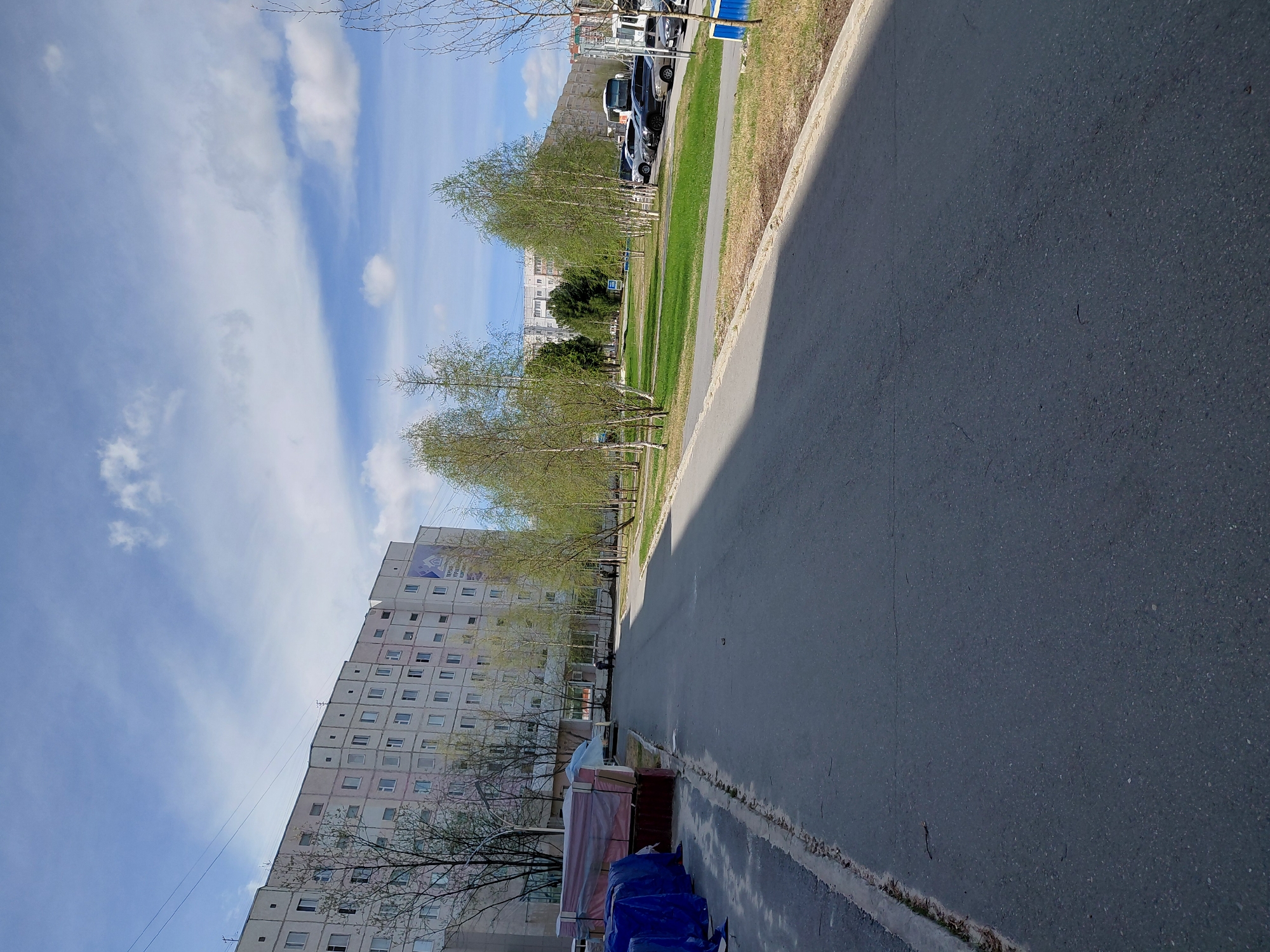 This media file captures a wide-angle view of a residential street in Nadym, Russia, on what appears to be a clear to partly cloudy spring day.
The scene is dominated by a large, multi-story apartment building on the left side of the frame, characteristic of post-Soviet architecture with numerous uniform windows and a light-colored facade. A blue and white banner with indistinct text and a logo is visible on the upper part of this building. In the foreground, below the apartment building, there are several items covered with blue tarps and lighter plastic sheeting, possibly construction materials or household belongings, with some wooden planks or crates also visible. A small figure, possibly a person walking or with a bicycle, is visible further down the street near the building.
Running from the foreground into the distance on the right is an asphalt road or wide paved path, bordered by a concrete curb and a strip of dry grass. Beyond this grassy strip, a line of deciduous trees with nascent, light green leaves stretches along the street, indicating early spring. More grassy areas are visible behind these trees, and several parked cars can be seen further down the road. In the far distance, another building is partially visible behind the trees and cars.
The sky is bright blue with scattered white clouds, some appearing wispy and others more defined. Shadows are cast across the asphalt path from the left, suggesting the sun is angled, possibly during the late morning or early afternoon. The overall impression is a typical, quiet street scene in a residential area.