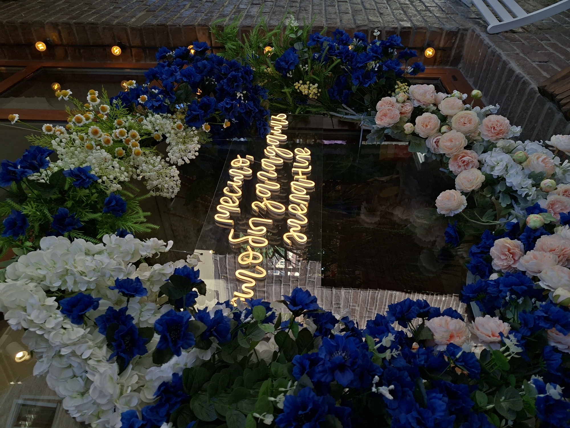 The image shows a storefront decorated with artificial flowers, likely in Estosadok, Russia. The storefront features a window with a neon sign that reads "Место, чтобы загадать желание" which translates to "A place to make a wish". The flowers are a mix of white, blue, and pink, with some smaller white and yellow flowers interspersed. The storefront is made of brick, and there are small yellow lights visible on the exterior. A white chair is partially visible on the right side of the image.