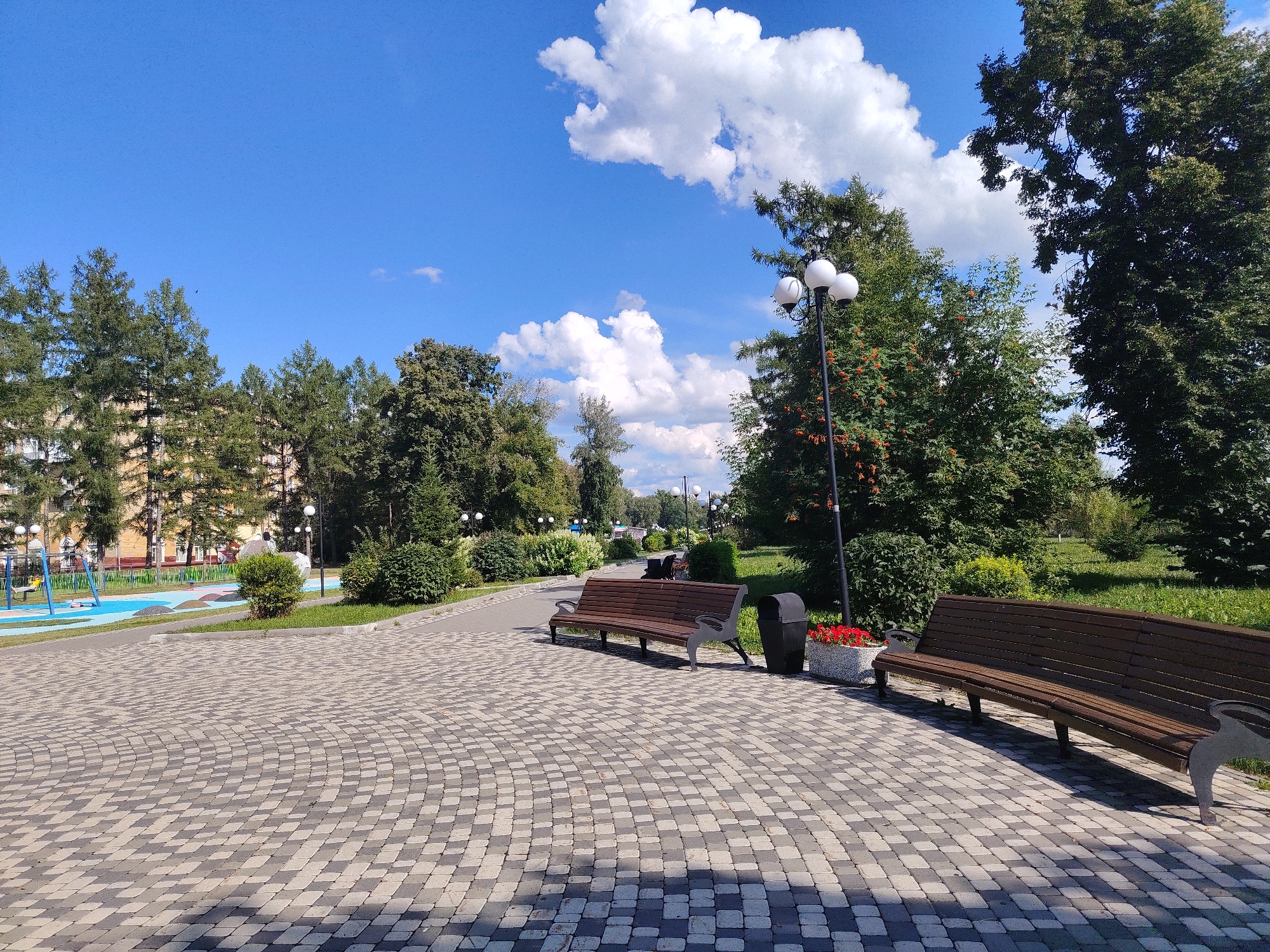The image depicts a public park in Kemerovo, Russia, on a sunny day with a clear blue sky and fluffy white clouds. In the foreground, a paved pathway made of gray and black interlocking stones curves to the right. Two wooden benches with ornate dark gray metal legs are placed on the pathway. To the right of the first bench is a dark gray trash receptacle. In front of the second bench, a rectangular stone planter holds bright red flowers.
The middle ground features a paved walkway bordered by green grass and trees. Several streetlights with multiple globes line the walkway. Beyond the walkway, a playground area with blue and yellow equipment is visible. Tall evergreen trees and deciduous trees with green foliage form a lush backdrop. In the far distance, a building with light-colored walls and windows can be seen behind the trees. The overall scene is peaceful and suggests a well-maintained public space.