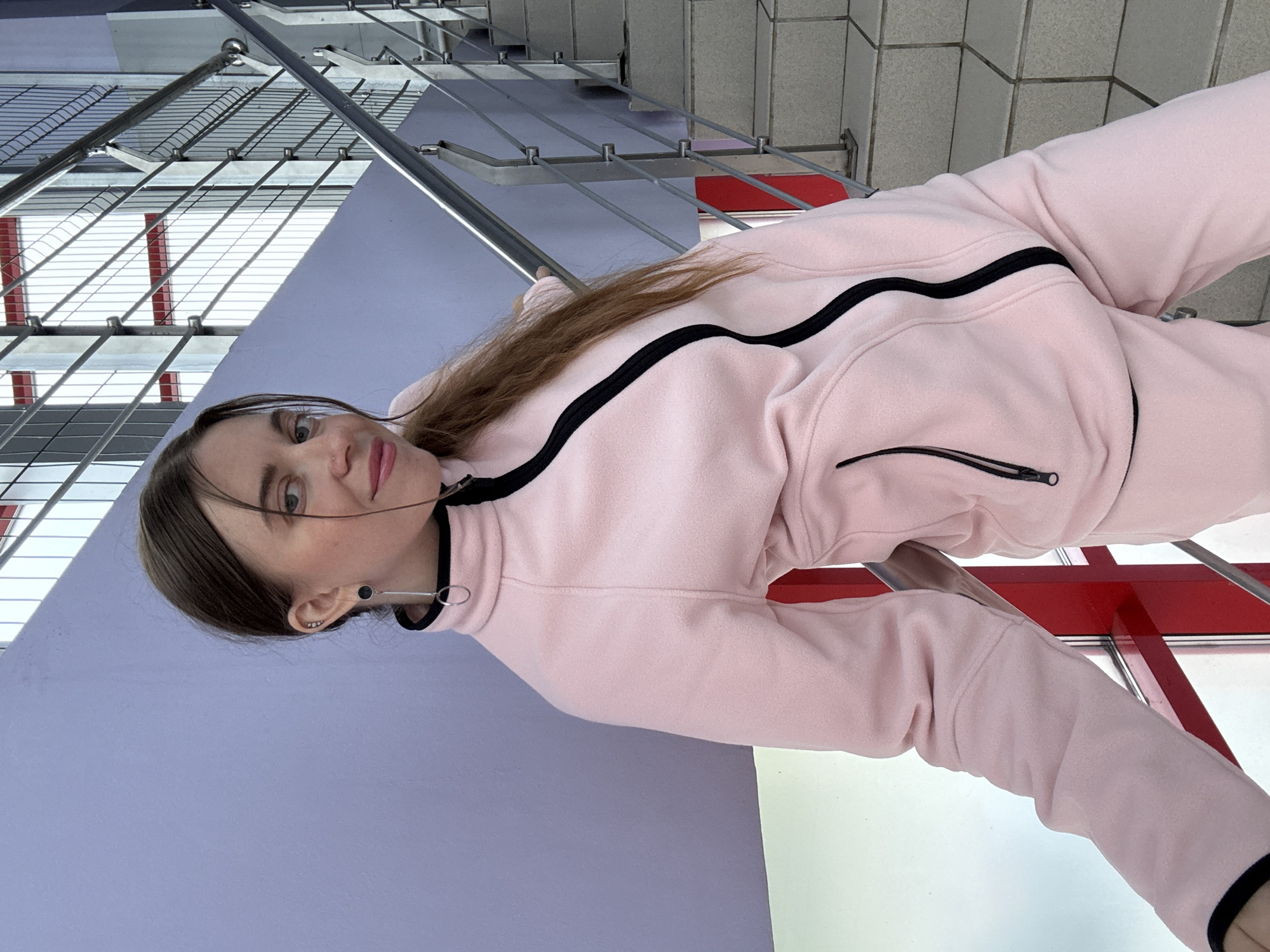 A young woman with long brown hair stands on a stairway, leaning against a purple wall. She is wearing a light pink tracksuit with a black zipper and trim. She has a silver earring in her left ear and a subtle nose ring. The stairway has metal railings and concrete steps. The background shows a metal fence and red accents on the structure.