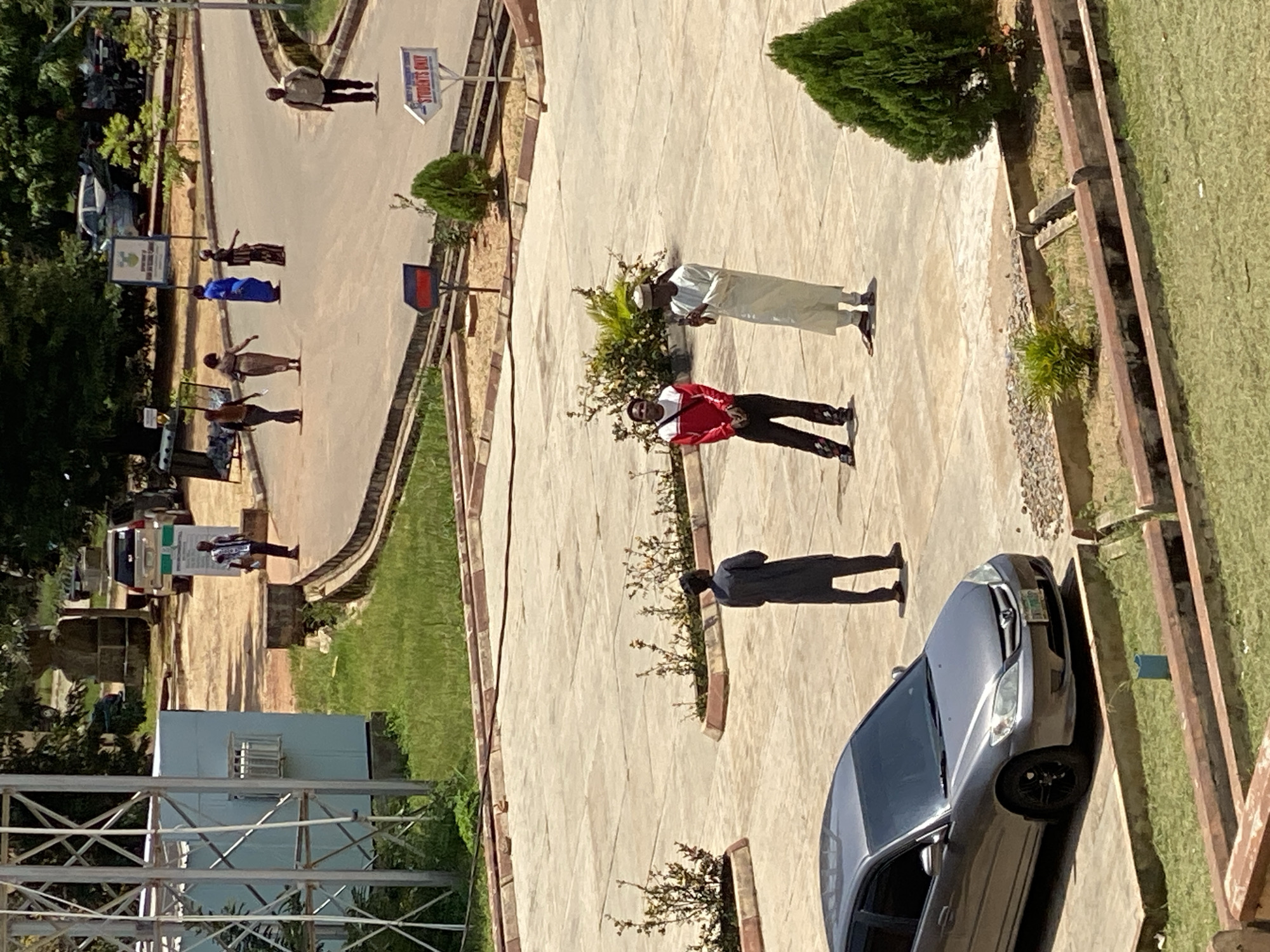 The image shows a group of people in an outdoor setting, possibly a campus or a public space, in Jos, Nigeria. The scene is bathed in bright sunlight, indicating it is daytime, and the weather appears to be clear.
There are several individuals visible. In the foreground, a person in a red jacket and dark pants is standing. Further back, another person in a white robe and a cap is standing. To their left, a person in a dark jacket is also standing. Scattered throughout the image are other individuals, some in traditional attire. One person appears to be walking, while others are standing still.
Objects in the scene include a dark grey car parked on the right side of the frame. There are also signs visible, one of which reads "STUDENTS ONLY". Trees and grassy areas suggest a well-maintained environment. A structure resembling a metal tower or scaffolding is present on the far left.
The people appear to be engaged in various activities, some standing and conversing, while others seem to be walking or waiting. The overall mood seems neutral, with no strong indicators of emotion. The architecture and landscaping suggest a location in a developed urban area. The presence of the "STUDENTS ONLY" sign strongly implies an educational institution.