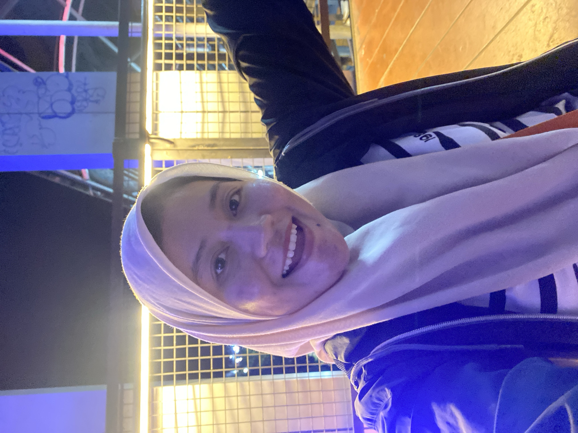 The image is a selfie taken at night in Banjarbaru, Indonesia, likely near a Ferris wheel or amusement park attraction, given the background structures with blue and pink lights and graffiti. The subject, a woman wearing a light-colored hijab and a dark jacket over a striped shirt, smiles broadly, her teeth visible. Her right arm is extended out of frame, suggesting she is holding the camera or posing. The lighting is artificial and casts a purple hue on her face. The foreground shows a wooden floor, likely part of an outdoor seating area or walkway.