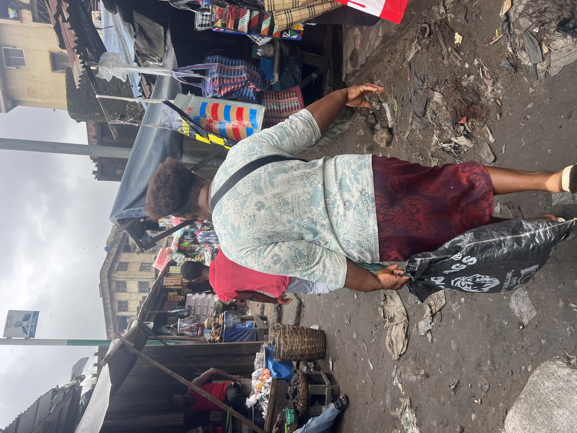 The image depicts an outdoor market scene in Nkpor, Nigeria. In the foreground, a woman walks away from the camera, carrying a black plastic bag in her right hand and wearing a light blue patterned shirt and a dark red skirt. She has an afro hairstyle and a black strap across her chest. Behind her, a person in a bright pink shirt walks in the same direction. Further back, a person in a red tank top is seated on a wooden structure. The market stalls are constructed with various materials, including corrugated metal and tarpaulin, with goods like produce and bags displayed. The ground is unpaved and appears dirty, with debris scattered around. In the background, multi-story buildings are visible. A lamppost with a political poster is also present. The sky is overcast.
