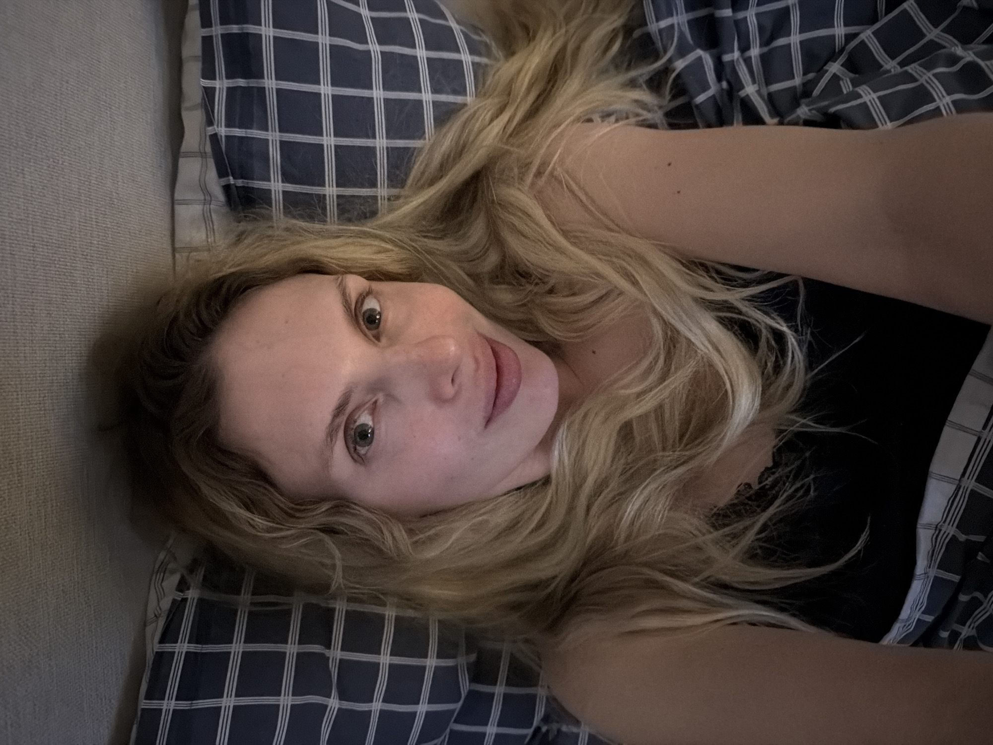 A blonde-haired woman lies on a bed. The bed has a grey and white plaid pillowcase. She is wearing a black shirt. The location is unknown, and there is no evidence to suggest it is in Budva, Montenegro.