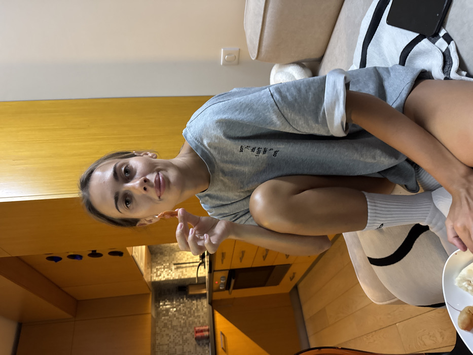 A young woman in a grey sweatshirt and white socks sits on a couch, facing the camera. She holds a small, orange object in her hand, possibly food. The kitchen is visible in the background with yellow cabinets and a mosaic backsplash. Part of a plate of food and a tablet are also in view. The overall impression is casual and domestic.