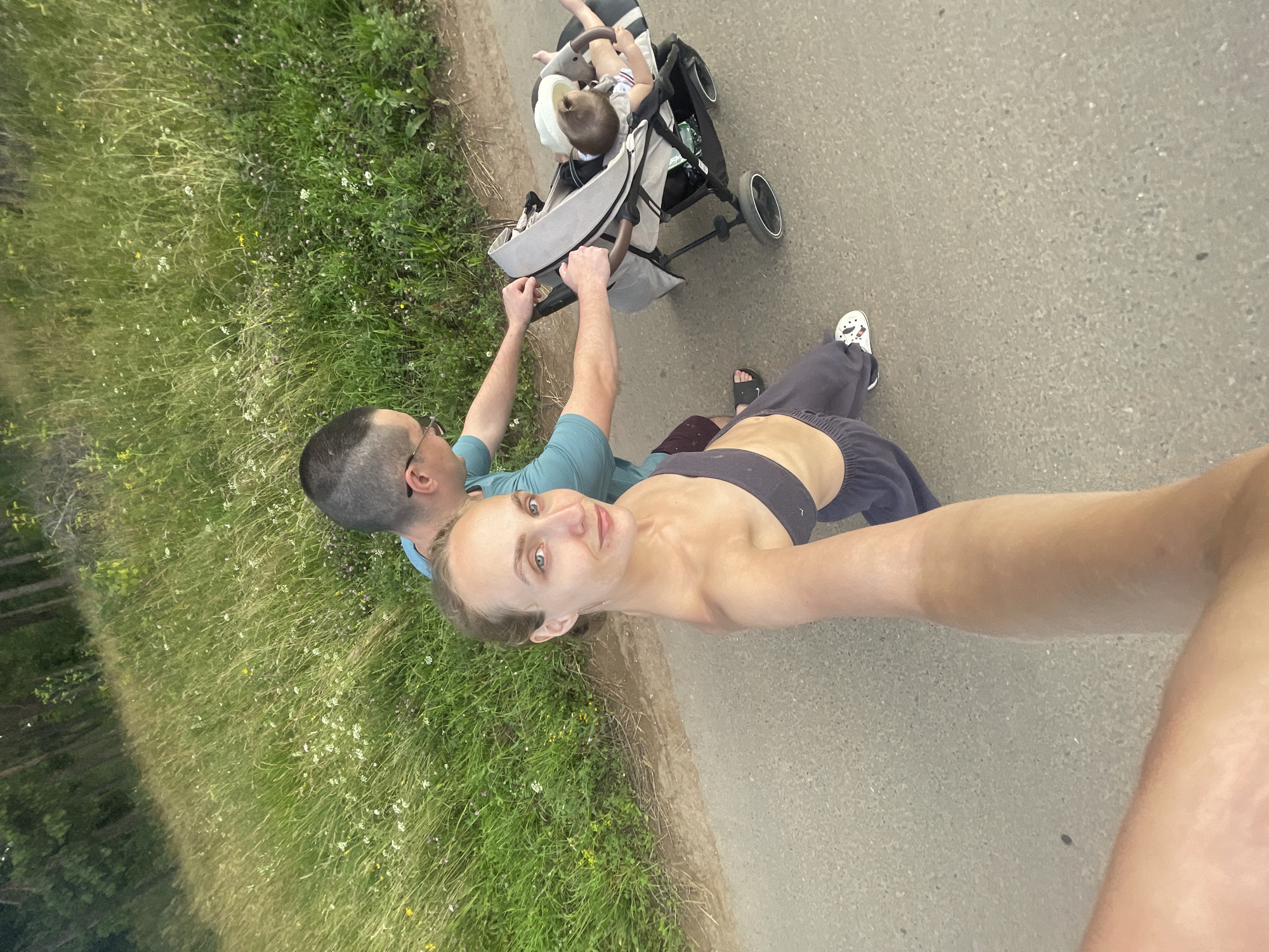A high-angle, wide shot captures a family on a paved path in a rural area, possibly near Podshivalovo, Russia, with a grassy field and trees to the left. A man in a teal t-shirt and burgundy shorts pushes a gray stroller, in which a baby wearing a white hat lies. To the left of the stroller, a woman in a dark grey sports bra and grey sweatpants, also facing the camera, is taking a selfie. She has blonde hair pulled back and is wearing white sneakers. The scene appears to be during the daytime, with natural light illuminating the subjects and the surrounding greenery.