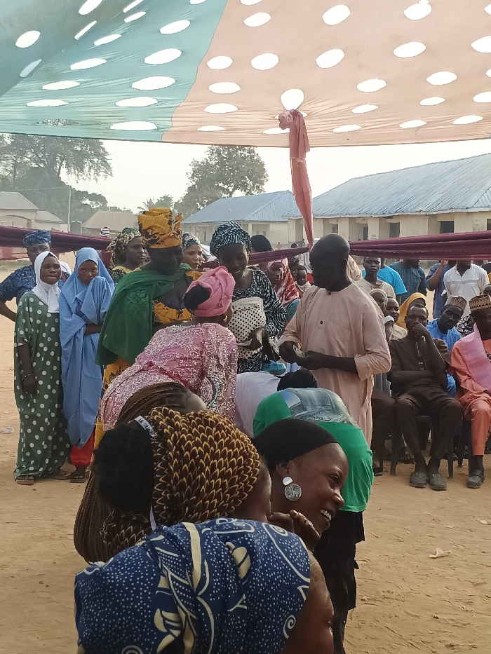 This outdoor image depicts a lively social gathering in Doma, Nigeria, under a large, temporary canopy. The canopy is made of fabric, featuring light green and beige sections with white polka dots, and is tied back in places.
Numerous people are present, a mix of adults, with many women dressed in colorful traditional African attire, including various head coverings and patterned dresses. In the center, a man in a light peach-colored tunic stands, appearing to interact with a woman who is bending forward in front of him. Other individuals surround them, observing the activity. Many faces show engagement, with some people smiling or laughing, particularly a woman in the foreground on the right wearing a black headscarf and distinct circular earrings. To the left, a group of women stands in what appears to be a line, some wearing hijabs.
In the background, additional attendees are seated on chairs. The ground beneath is bare earth or sand. Simple, single-story buildings with light-colored walls and corrugated metal roofs are visible in the distance, interspersed with trees, indicating a rural or semi-urban setting. The natural light suggests it is daytime, with no harsh shadows implying midday, possibly morning or late afternoon. The overall atmosphere appears communal and celebratory. There is no visible text in the image.