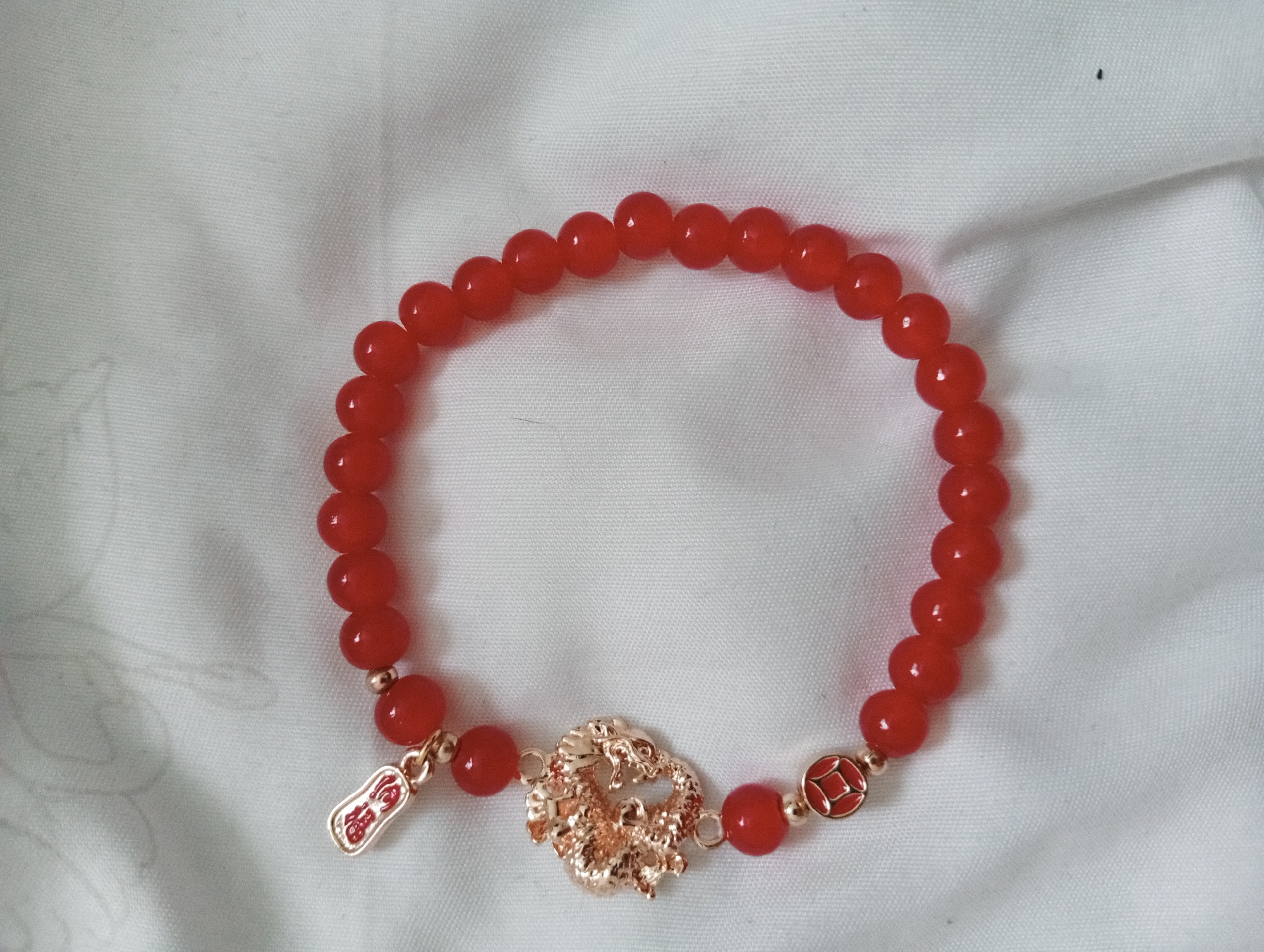 A red bead bracelet rests on a white fabric surface. The bracelet consists of round, reddish-orange beads strung together. The bracelet is circular, but it is not closed and is instead open like an incomplete circle. At the bottom of the bracelet, there is a golden charm in the shape of a dragon, attached between two beads with gold colored accents. To the left of the dragon charm is a smaller red and gold charm in a rectangular shape. There is a small black speck visible at the top of the frame. The scene appears to be a close-up shot.