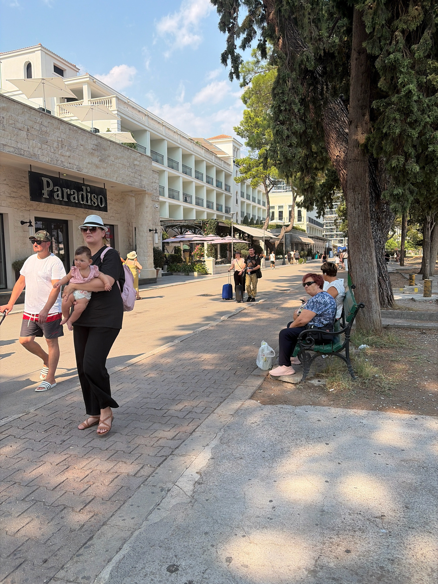 The image depicts a sunny daytime scene on a paved walkway lined with trees and buildings. In the foreground, a woman in black pants and a black shirt, wearing a white hat and sunglasses, holds a baby wearing a pink top and a diaper. She is walking towards the viewer, with her left foot slightly forward. To her left, a man in a white t-shirt, gray shorts with a red and white waistband, a camouflage cap, and sunglasses is walking away from the viewer, pushing a stroller with his left hand. He is wearing white slide sandals. Behind them, on the right side of the walkway, two women are sitting on a dark green park bench. The woman closer to the foreground is wearing a blue and white patterned shirt, dark pants, sunglasses, and pink sneakers. The woman behind her is wearing a white top and dark pants. A clear plastic bag is on the ground to the left of the bench.
The background features a modern building with multiple balconies and white railings, and a sign above an entrance reads "Paradiso". Further down the walkway, other pedestrians are visible, some carrying luggage, and outdoor seating areas with umbrellas are set up. Trees with thick trunks and dense foliage line the right side of the walkway, casting shadows on the ground. The ground surface is a mixture of interlocking pavers and concrete. The sky above is blue with scattered white clouds. The scene appears to be a promenade or street in a resort area, possibly in Budva, Montenegro, given the architectural style and the presence of a beach-like environment.