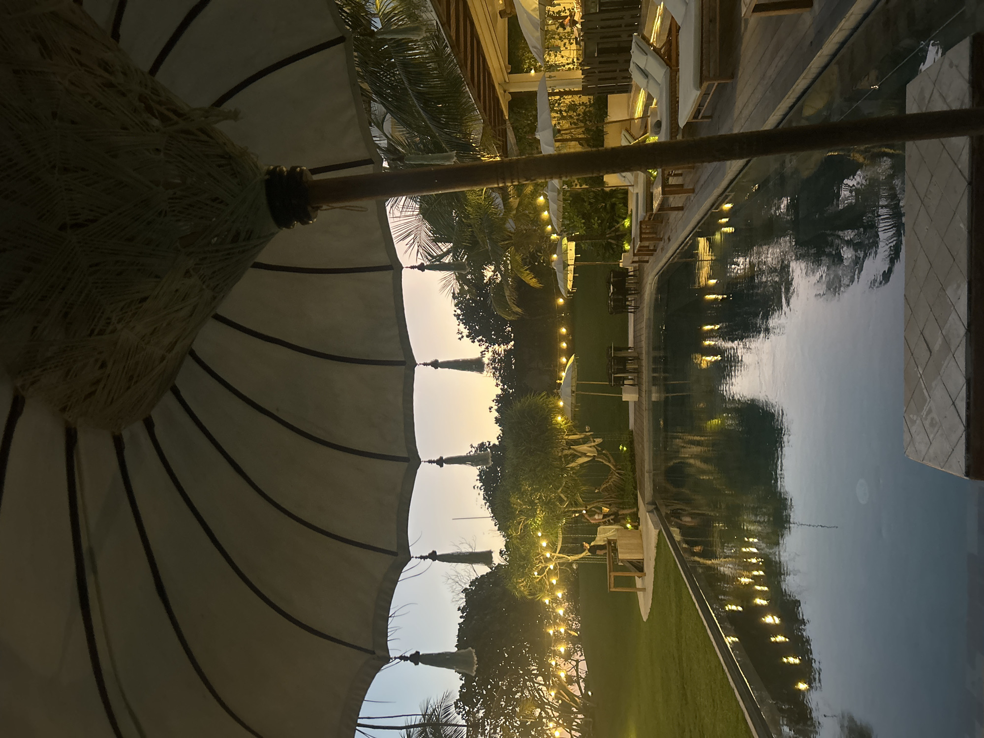This is a medium shot photograph taken at dusk, likely at a resort or hotel, located in Kediri, Indonesia. The scene is centered around a swimming pool, with lounge chairs and umbrellas positioned on a wooden deck adjacent to it. The pool water reflects the surrounding lights and palm trees. In the foreground, a large umbrella partially obscures the view, with decorative tassels hanging from its edge. The background features lush greenery, trees adorned with string lights, and a hint of buildings with awnings. The sky displays a gradient of warm sunset colors. Two individuals are visible near a table under one of the lit trees, one appearing to be interacting with a counter or service area. The overall atmosphere is tranquil and inviting, suggesting an evening leisure setting.
