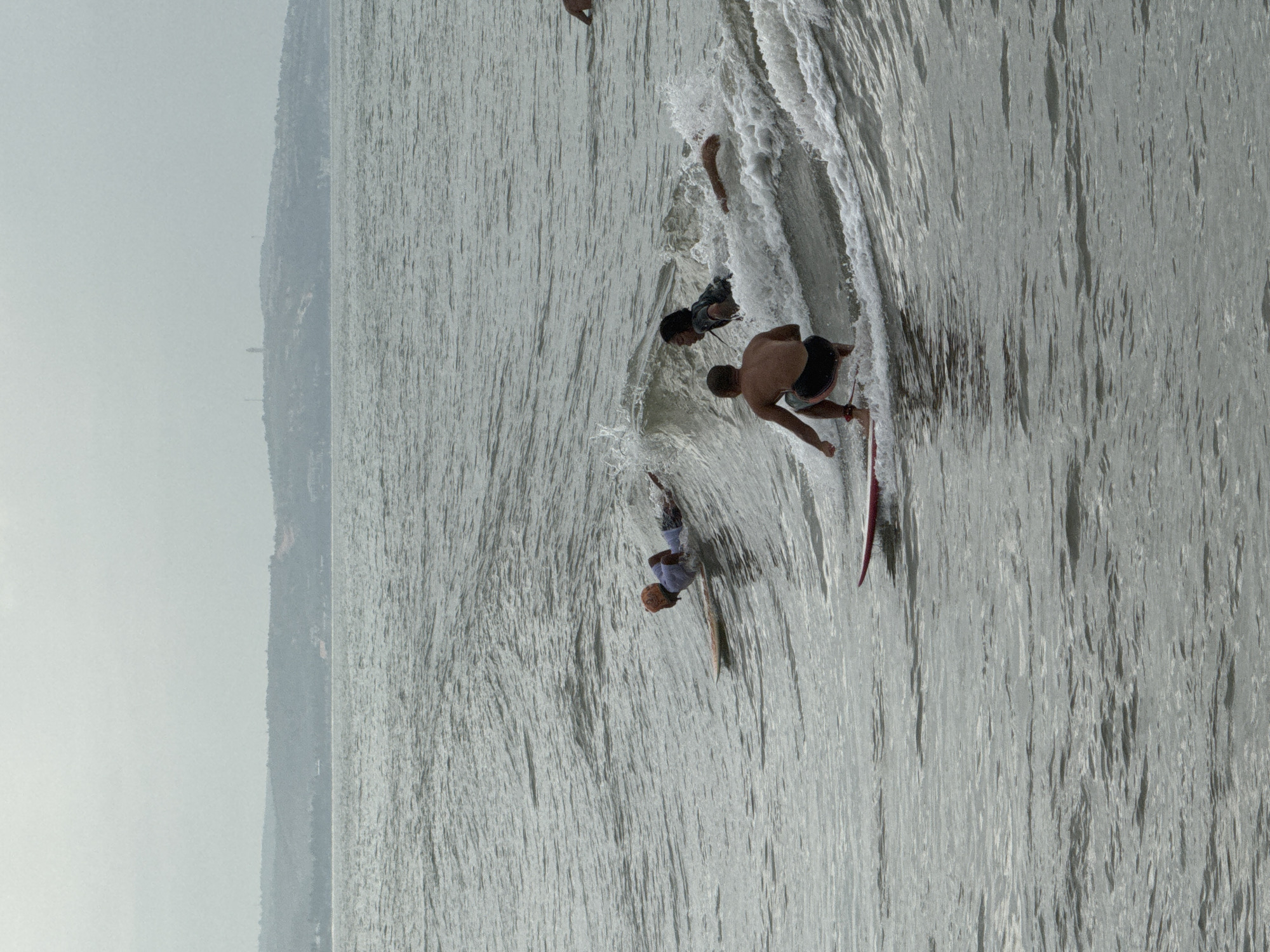 Four individuals are surfing on a choppy ocean under an overcast sky. The middle ground features three surfers actively riding waves. In the foreground, one surfer is positioned on a red surfboard, crouched low and riding a wave. To the left and slightly behind, another surfer lies prone on a light-colored surfboard. Further back and to the right, a third surfer is partially submerged in a wave, with only their head and shoulders visible. A fourth individual, further in the distance on the right, is partially visible, also in the water. The background consists of a hazy shoreline with hills and vegetation. This scene is likely located in the waters off the coast of Phan Thiết, Vietnam, a region known for its surfing conditions.