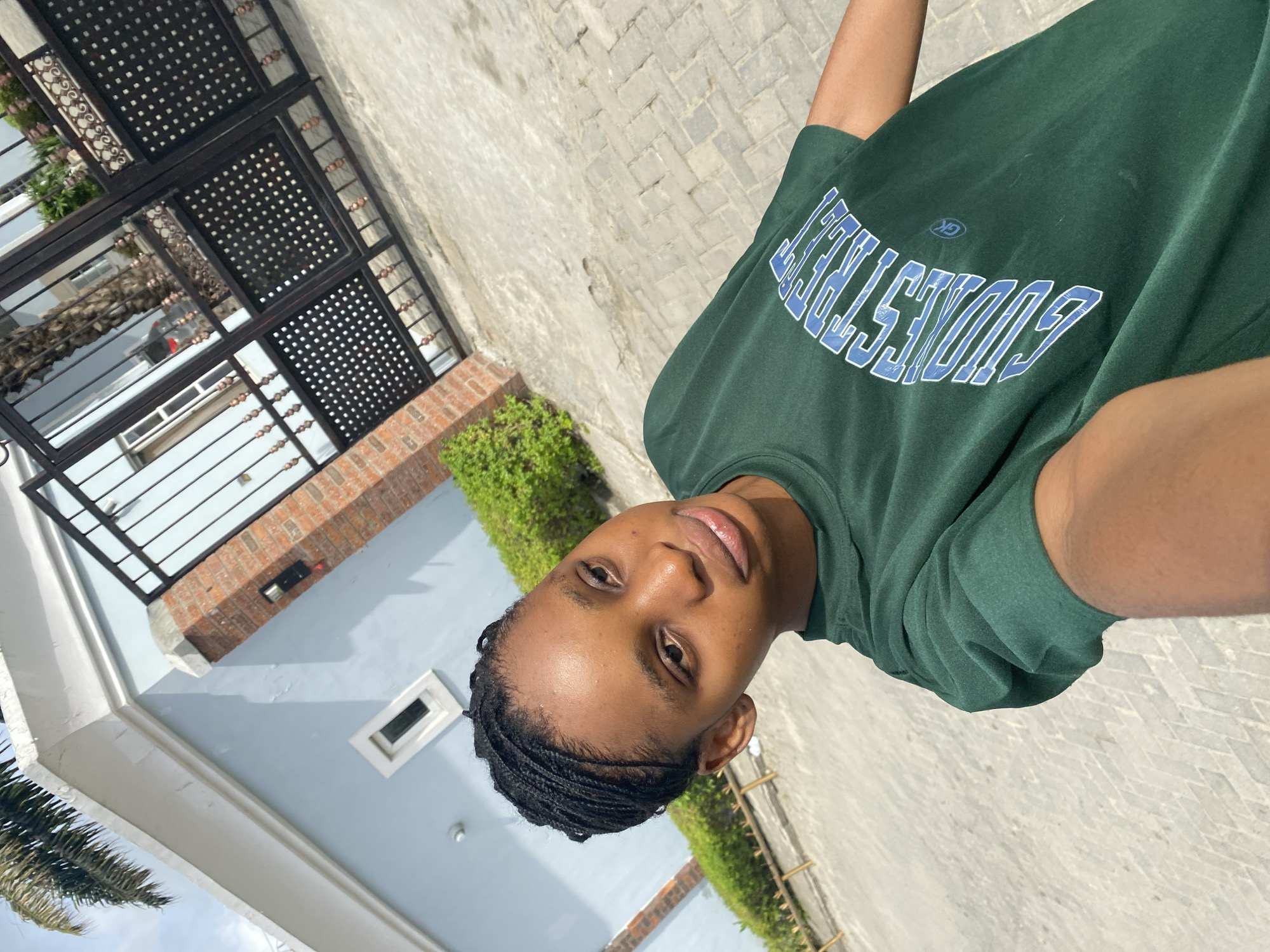 A person with braided hair is wearing a green shirt with blue lettering. They are standing outside near a building and pavement. The building is light blue with a brick detail. There is greenery present. The location is possibly Iranla, Nigeria.