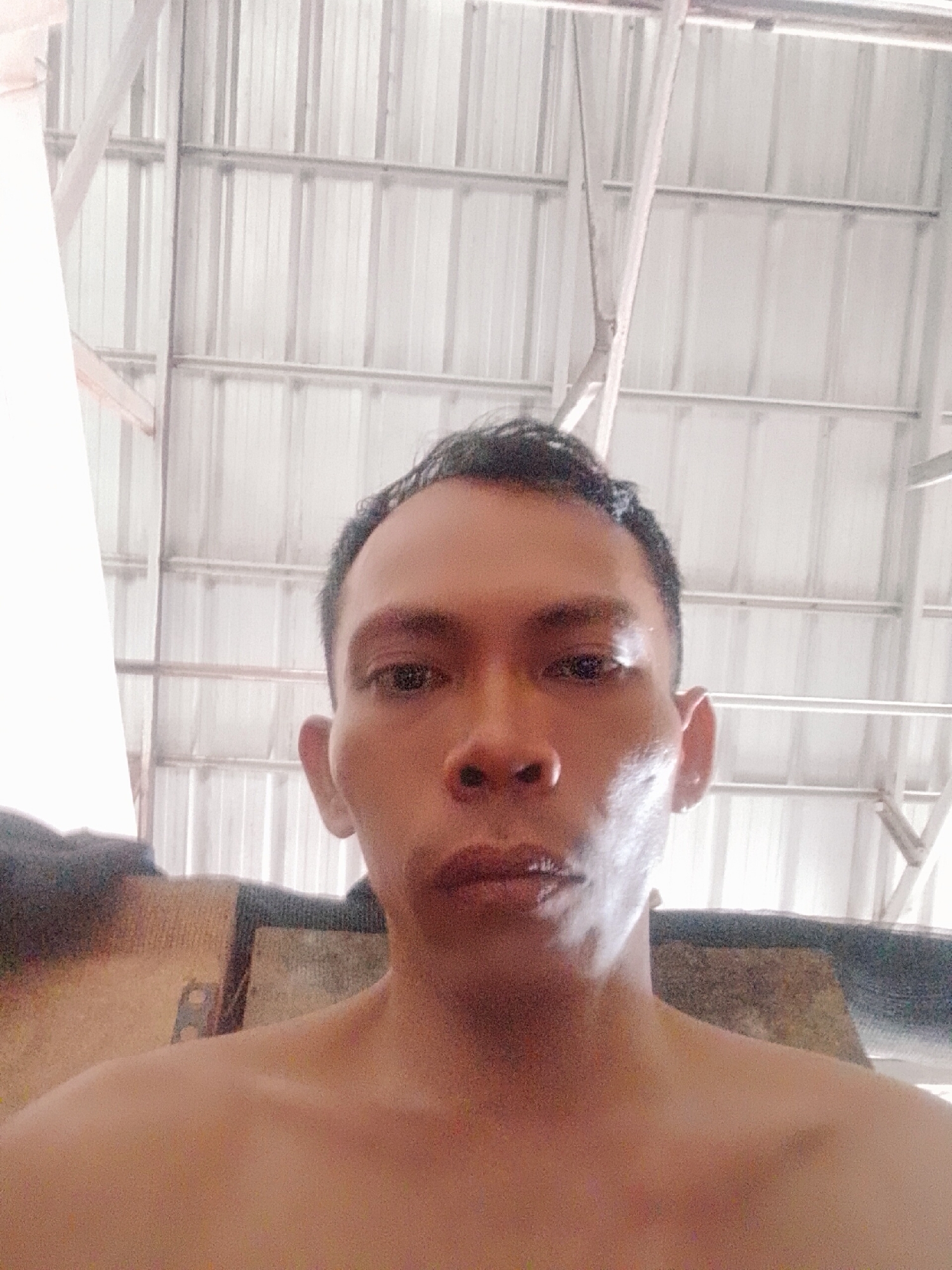 A head-and-shoulders shot of a man with short black hair. The man has brown eyes and a serious expression. He is bare-chested. The background shows a metal ceiling with a support structure. Based on the location information, the setting is potentially in Bukit Ketok, Indonesia.