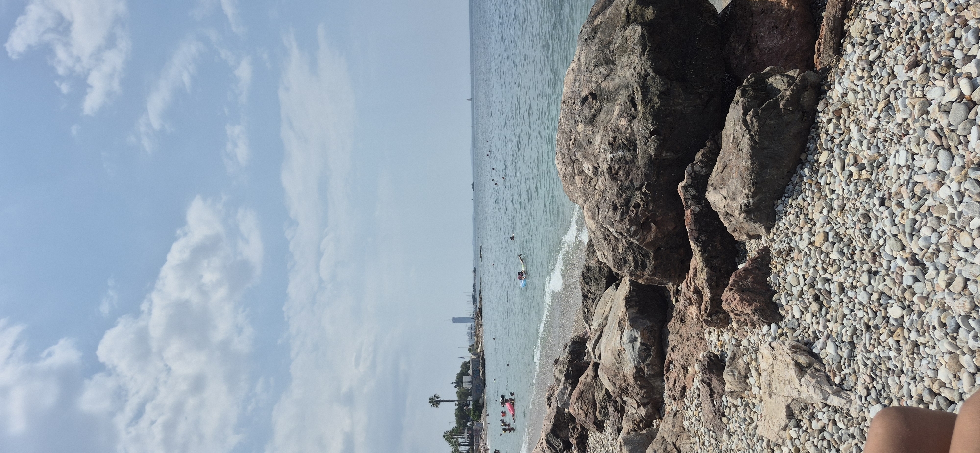 The image depicts a rocky beach in Paleo Faliro, Greece, with people swimming and relaxing in the calm, blue sea under a partly cloudy sky. In the foreground, large, weathered rocks and smooth pebbles cover the shoreline. The middle ground shows several individuals enjoying the water, some on inflatable rings. In the distance, a linear cityscape with buildings, including a prominent skyscraper, lines the coast. Palm trees are visible along the shore, and a few boats are scattered across the horizon. The overall scene is one of a typical summer day at a Mediterranean beach.