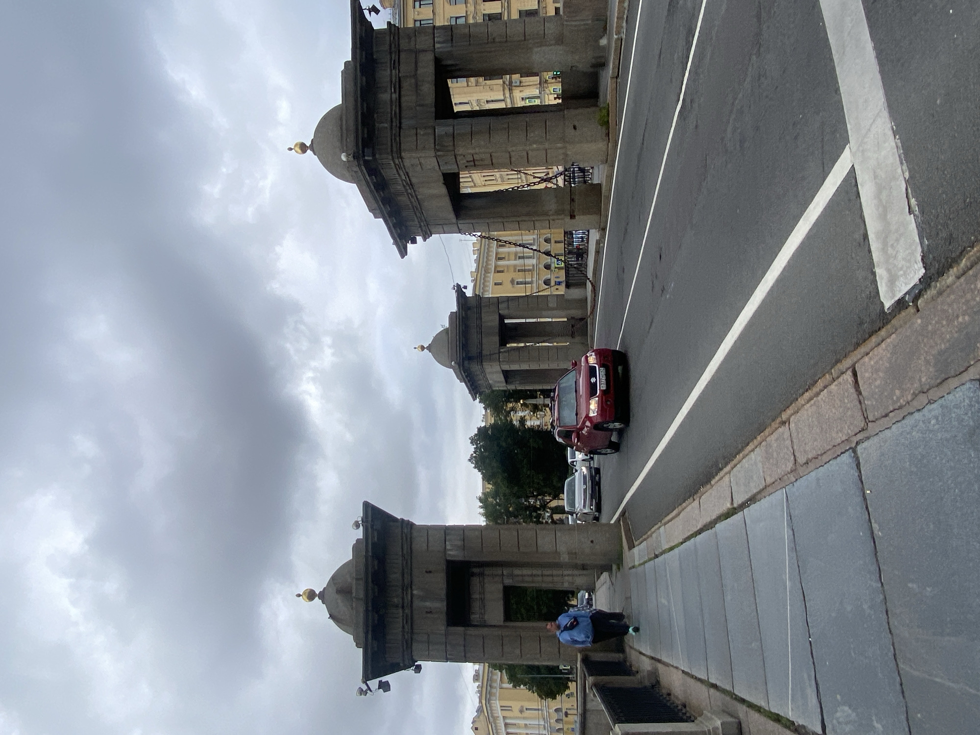 The image depicts a view of the Annunciation Bridge (also known as Lieutenant Schmidt Bridge) in Saint Petersburg, Russia, on a cloudy day. In the foreground, a pedestrian walkway is paved with large, dark grey stone slabs, some of which are cracked or misaligned. A person in a blue jacket and dark pants is walking away from the camera on the left side of the walkway, with a black railing to their left.
The bridge deck carries traffic. A red Suzuki SUV is in the right lane, moving away from the camera. Further ahead, a silver car and a white car are also visible. The road surface is asphalt with white lane markings.
The bridge is characterized by its distinctive stone pylons, two of which are prominent in the image. These pylons are square, massive structures with arched openings, topped with domes crowned with golden finials. Heavy chains are suspended between the pylons, suggesting a historical drawbridge mechanism.
In the background, classical architecture with yellow facades and numerous windows lines the embankment. Trees with green foliage are visible between the pylons and the buildings. The sky is overcast with grey clouds.