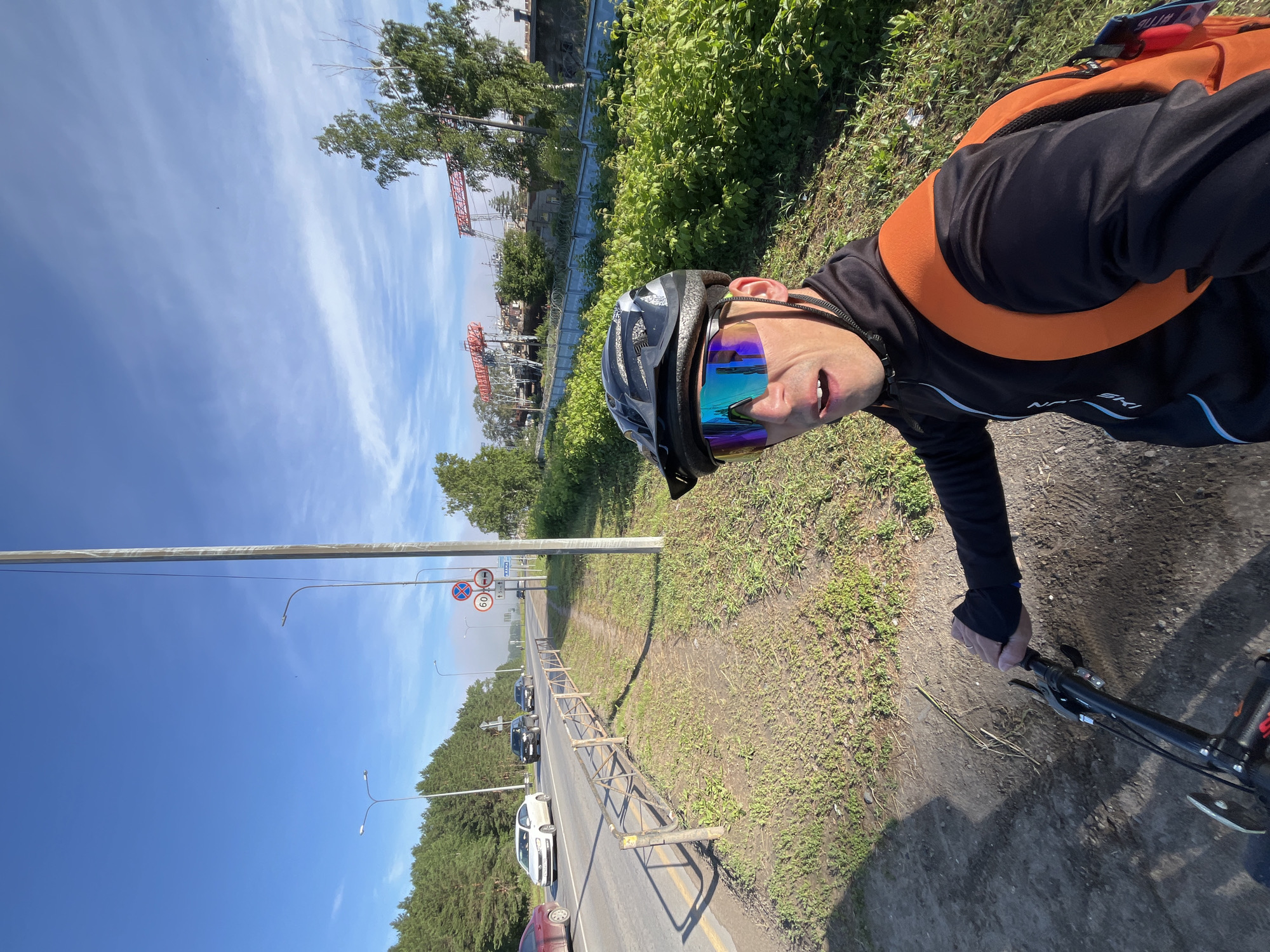 A first-person perspective selfie shows a man wearing a black cycling helmet and mirrored cycling glasses, with his mouth open, as he rides a bicycle. He is wearing a black cycling jacket with blue and white accents and an orange backpack. The man is on a dirt path next to a paved road. On the road, several cars are driving. Traffic signs are visible, including a 60 km/h speed limit sign and a no entry sign. In the background, there are trees, and further in the distance, industrial structures with red cranes are visible. The sky is blue with some wispy clouds. The scene appears to be on the outskirts of the city of Chaikovsky, Russia, possibly near an industrial area.