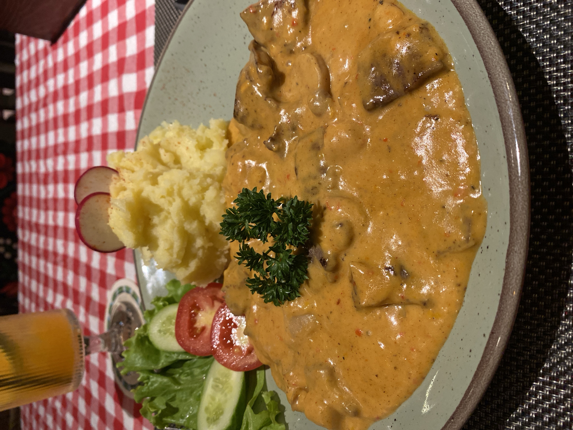 A close-up shot of a plate with a meal, seemingly taken indoors in Phu Thuy Ward, Vietnam, based on the context of the scene. The main dish appears to be meat, possibly beef stroganoff or a similar dish, covered in a creamy, yellowish-orange sauce with visible bits of pepper or spice. Accompanying the dish is a scoop of mashed potatoes garnished with a sprig of parsley, and a small side salad with lettuce, sliced cucumbers, sliced tomatoes, and a couple of radish slices. The plate has a greenish-blue hue with a brown rim and rests on a dark, textured surface, likely a placemat. A checkered red and white tablecloth is visible in the background, and a glass with a yellow drink is partially visible in the lower left corner of the frame.