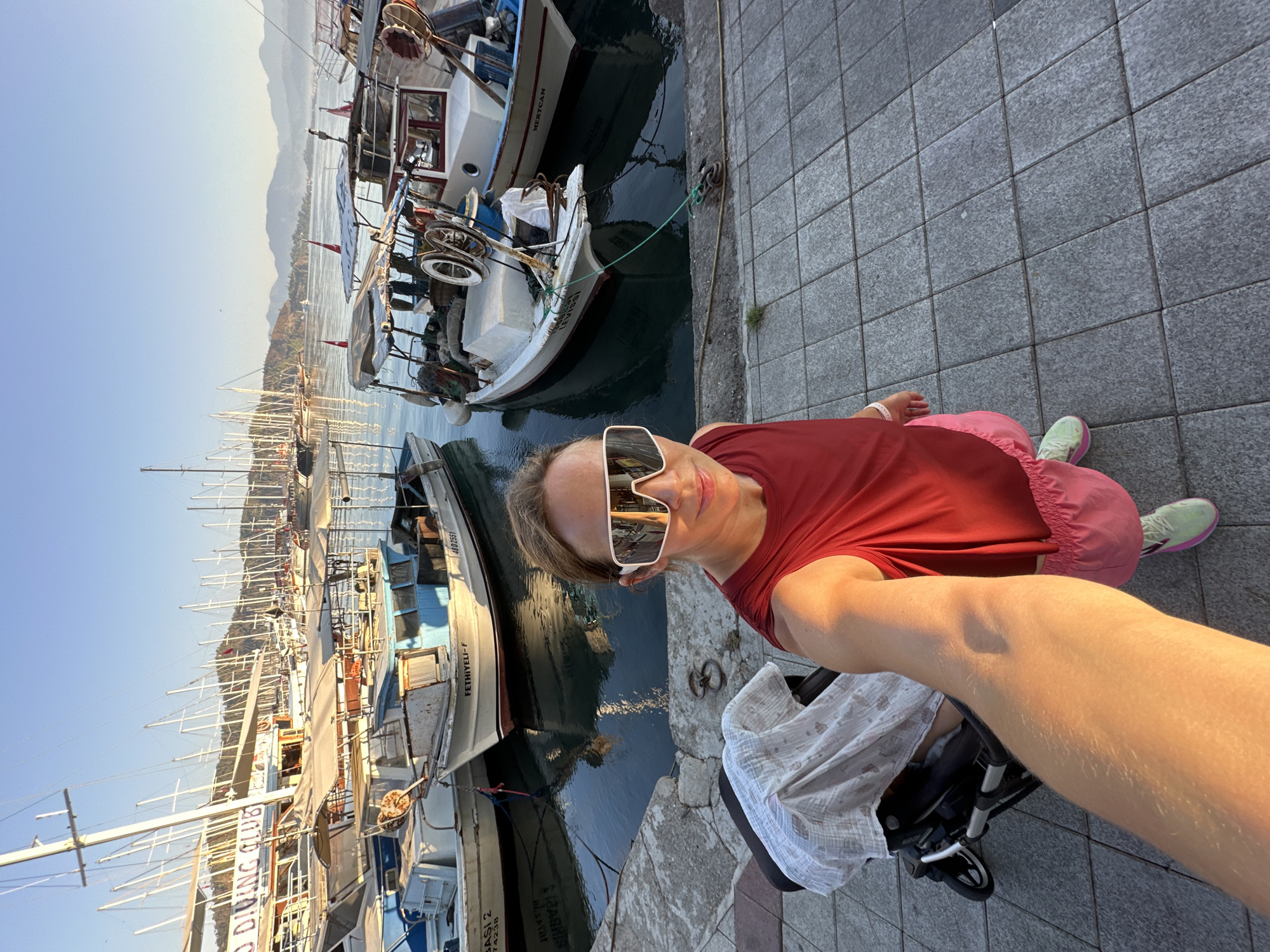 The image shows a woman taking a selfie in the harbor of Fethiye, Turkey. She is wearing large white sunglasses, a red sleeveless top, and pink shorts. She is standing on a paved walkway next to a stroller covered with a white blanket. Behind her, several boats are docked in the water, with masts and sails visible in the background. The sky is clear and blue, and the sun is casting a warm glow on the scene. The boats in the background are a mix of fishing boats and sailboats. The water is calm and reflects the boats and the sky. In the distance, mountains can be seen. The overall scene suggests a peaceful morning or afternoon in a coastal town.