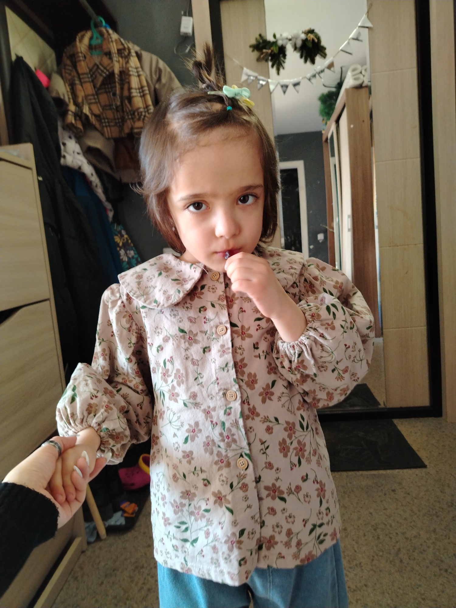 A young girl stands indoors, holding a purple object near her mouth. She has brown hair with a small ponytail held by a blue and green clip, and she is wearing a floral-patterned blouse with a large collar and gathered sleeves, paired with a blue skirt or pants. She looks directly at the camera with a somewhat apprehensive expression. A hand holds hers, likely that of an adult, with a black sleeve visible.
The background includes a closet with various clothes hanging inside and a mirror reflecting a portion of the interior space, including what appears to be holiday decorations. The location appears to be an apartment, with beige walls and a tiled floor. It is not possible to determine whether the location is in Noviy, Russia, or any other specific city based on the visual information provided.