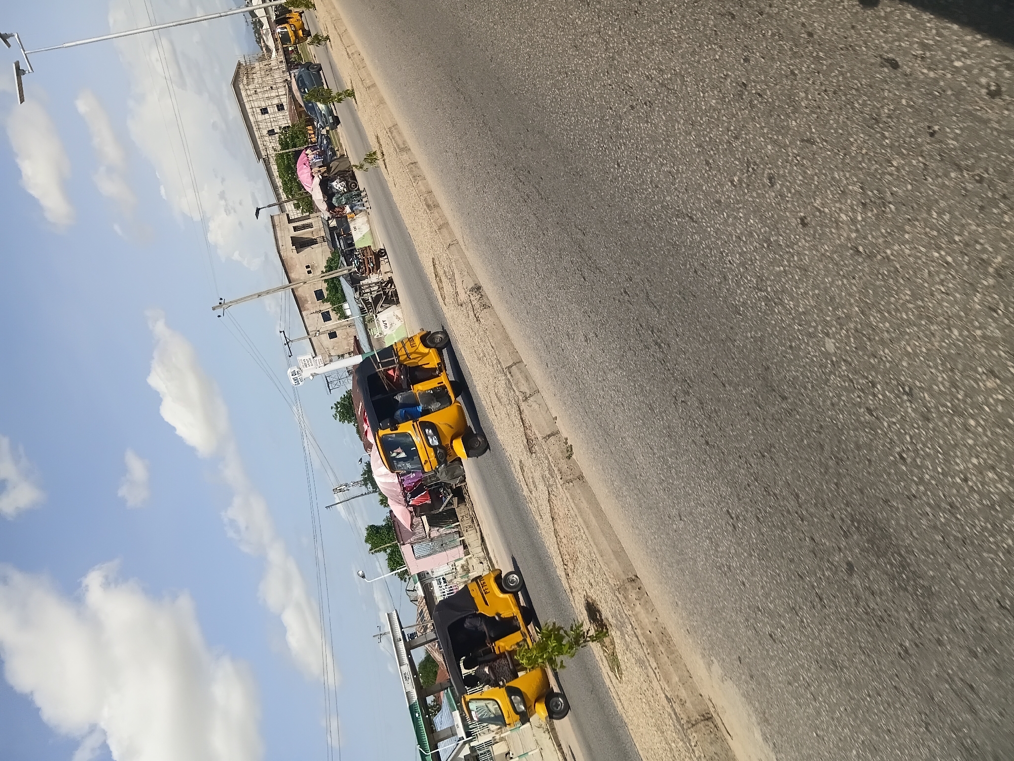 The image depicts a street scene in Jalingo, Nigeria, under a bright sky with scattered clouds. In the foreground, a paved road occupies the majority of the frame, with a textured surface. A sandy embankment with sparse vegetation borders the road.
Several yellow auto-rickshaws, also known as Keke Napeps, are present. Two are prominently visible on the road, each with passengers. One auto-rickshaw in the left midground has its black canopy up and appears to be carrying three individuals. Another auto-rickshaw further down the road on the right also has its canopy up and is carrying at least two people.
In the background, there are buildings, including what appears to be a petrol station with its canopy and pumps. Other structures are visible, some appearing unfinished or under construction. Various market stalls with colorful umbrellas are set up along the roadside, with people gathered around them. A silver car is also seen on the road. Utility poles and overhead wires are visible, indicating the presence of infrastructure. The overall impression is a bustling urban environment typical of many Nigerian cities.