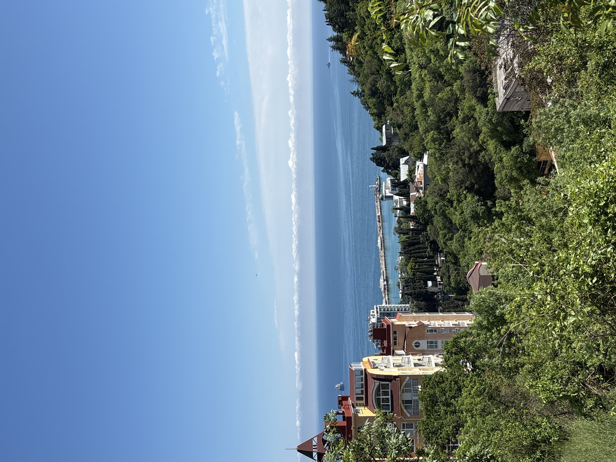 A slightly high-angle, outdoor shot faces the horizon from град Ялта, Ukraine. In the foreground are several buildings, colored beige, yellow, and red, partially obscured by verdant trees and shrubbery. Past the buildings, a narrow harbor is visible with a pier extending into the sea. Several boats are docked at the pier. The sea is a deep blue, contrasting with the bright blue sky. Streaks of white clouds are present in the sky, creating a linear pattern along the horizon. On the right, the coastline is densely covered with green trees.
