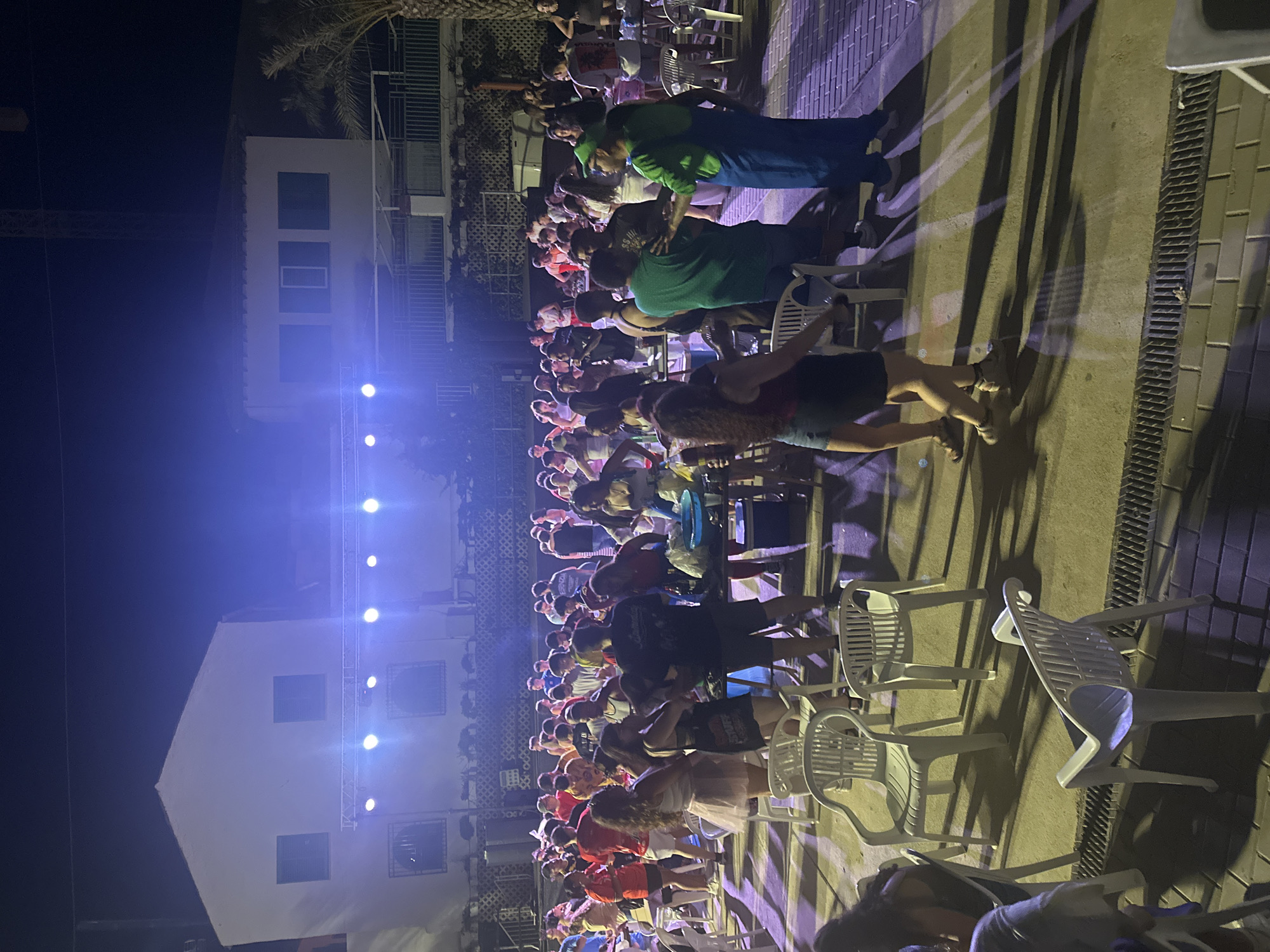 This image depicts a nighttime outdoor gathering, possibly a party or event, in Blanca, Spain. A crowd of approximately 30-40 people, mostly young adults, are present. The environment appears to be an open area with a paved ground, illuminated by stage lights that cast long shadows. In the background, there are buildings that suggest a residential or commercial setting. Several white plastic chairs are scattered around the area, some occupied and others empty. A makeshift table with drinks and other items is visible on the right side of the frame. One individual in the foreground is dressed in a green outfit with blue overalls, resembling a character costume. People are standing, conversing, and some appear to be dancing, suggesting a lively atmosphere. The overall scene is indicative of a social event taking place after dark.