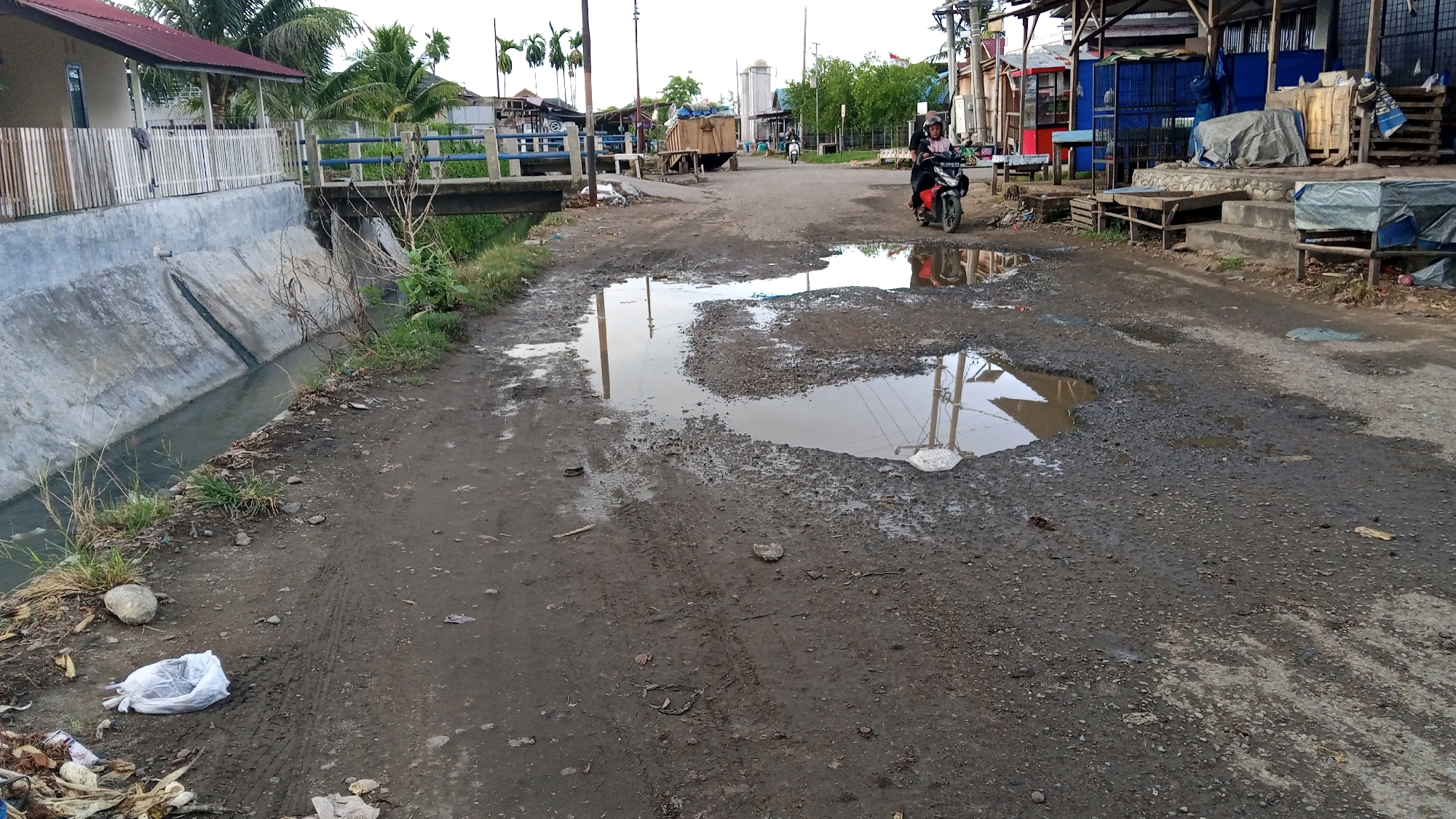 The image depicts a rural street scene in Bireuen, Indonesia, characterized by a poorly maintained, muddy road with large puddles. A person is riding a black motorcycle with a pink scarf and helmet. The road is bordered on the left by a concrete canal with water, and on the right by a row of small shops and buildings constructed with wood and corrugated metal roofing. Debris, including plastic bags and other trash, is scattered along the roadside. In the background, a truck is parked on the road, and several palm trees are visible. The overall condition of the road suggests a lack of infrastructure development.