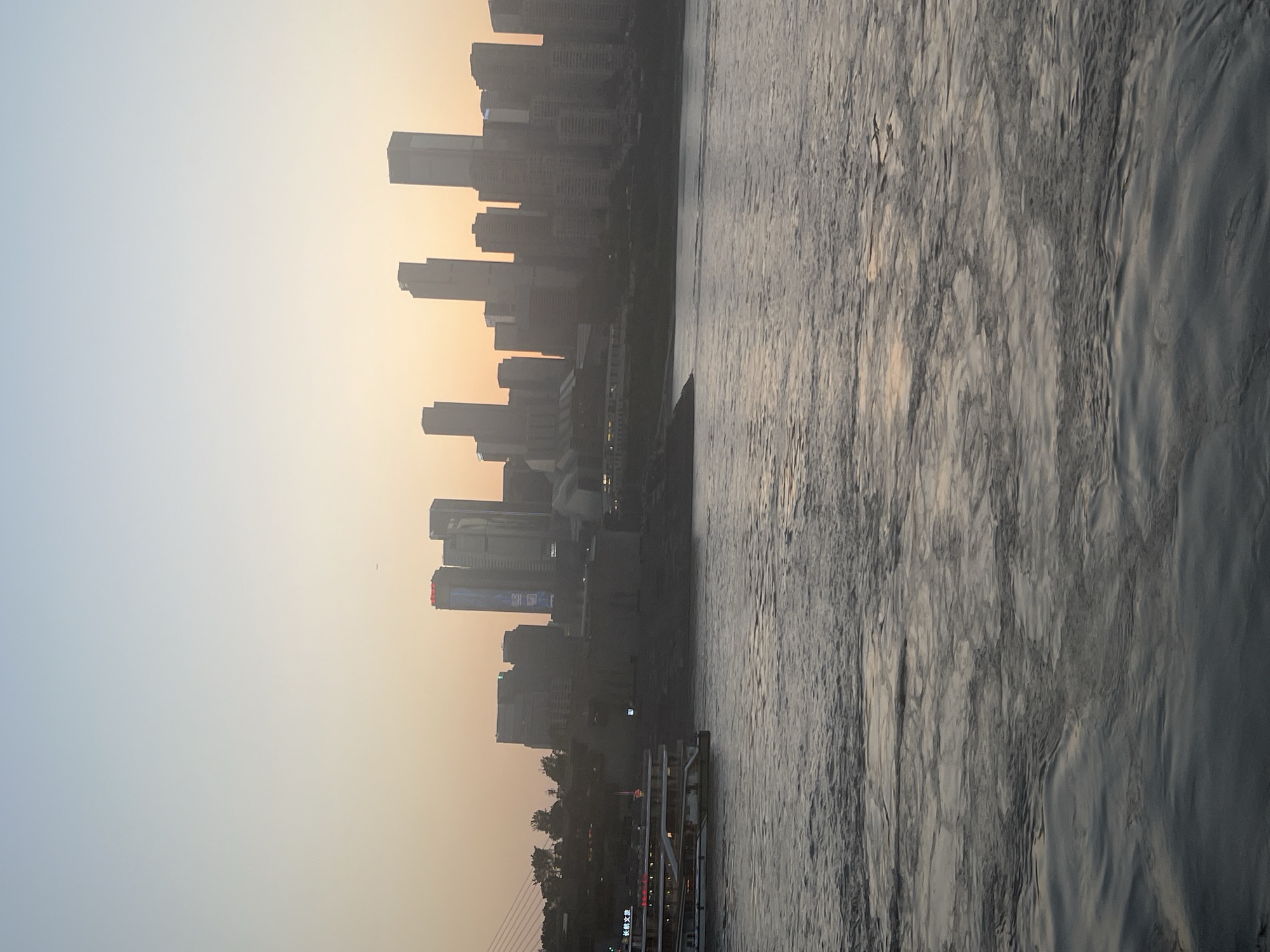 A hazy sunset casts a warm glow over a cityscape of tall buildings in Nan'an District, China. In the foreground, a wide river flows with gentle ripples, reflecting the soft light. On the left side of the river, a multi-level tourist boat with illuminated windows is docked along a waterfront. The buildings in the background rise dramatically, their forms softened by the atmospheric haze. A few bare trees are visible on the riverbank. The sky transitions from a pale blue at the top to a peachy orange near the horizon.
