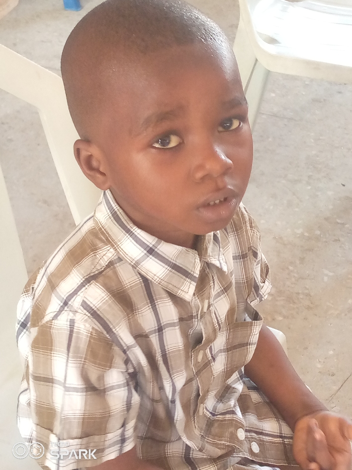 The image depicts a young, dark-skinned male child positioned centrally, looking directly forward. The child has short, dark, closely cropped hair. His eyes are open and directed towards the viewer, with a slightly open mouth revealing the upper central incisors. He is dressed in a short-sleeved collared shirt featuring a plaid pattern in shades of brown, white, and faint blue/gray, secured by white buttons down the front. The right forearm and hand are partially visible in the lower right quadrant of the image, appearing to rest on his lap. The background is out of focus, revealing a light-colored floor and portions of a white plastic chair to the left and upper right of the subject. The scene is illuminated by ambient, diffuse light. The photograph was captured in Ajaokuta, Nigeria. A 'TECNO SPARK' watermark is visible in the bottom-left corner.