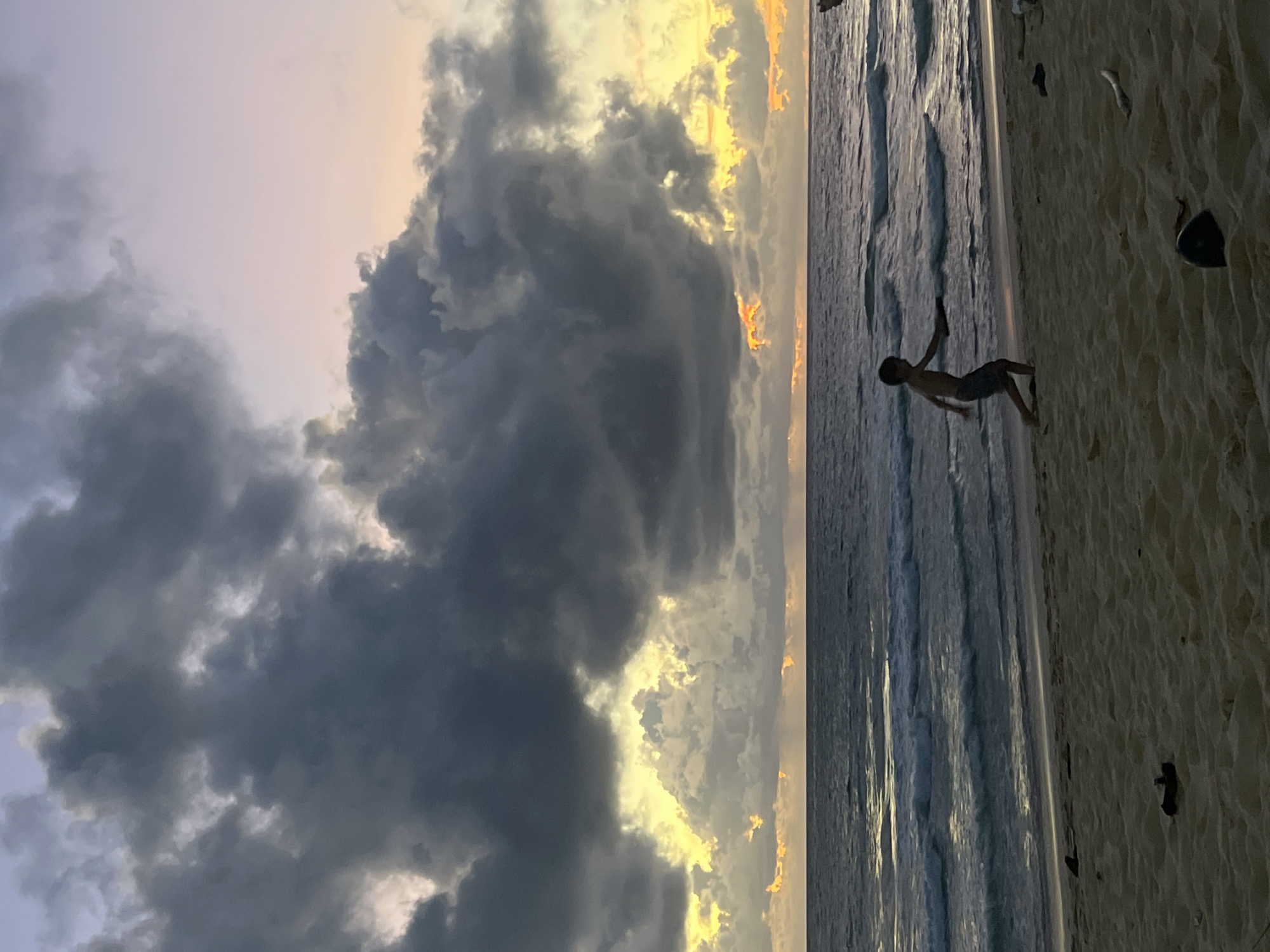 A boy is playing on a sandy beach with the ocean in the background. The sky is filled with dramatic, dark clouds that are lit from behind by the setting sun, creating a yellow and orange glow. The boy is running and appears to be holding a frisbee or a similar object. There are some pieces of driftwood and debris scattered on the sand. The water is dark and shows waves rolling towards the shore. The scene suggests a late afternoon or early evening setting. The location is not specified in the image or the prompt beyond a general geographic area.